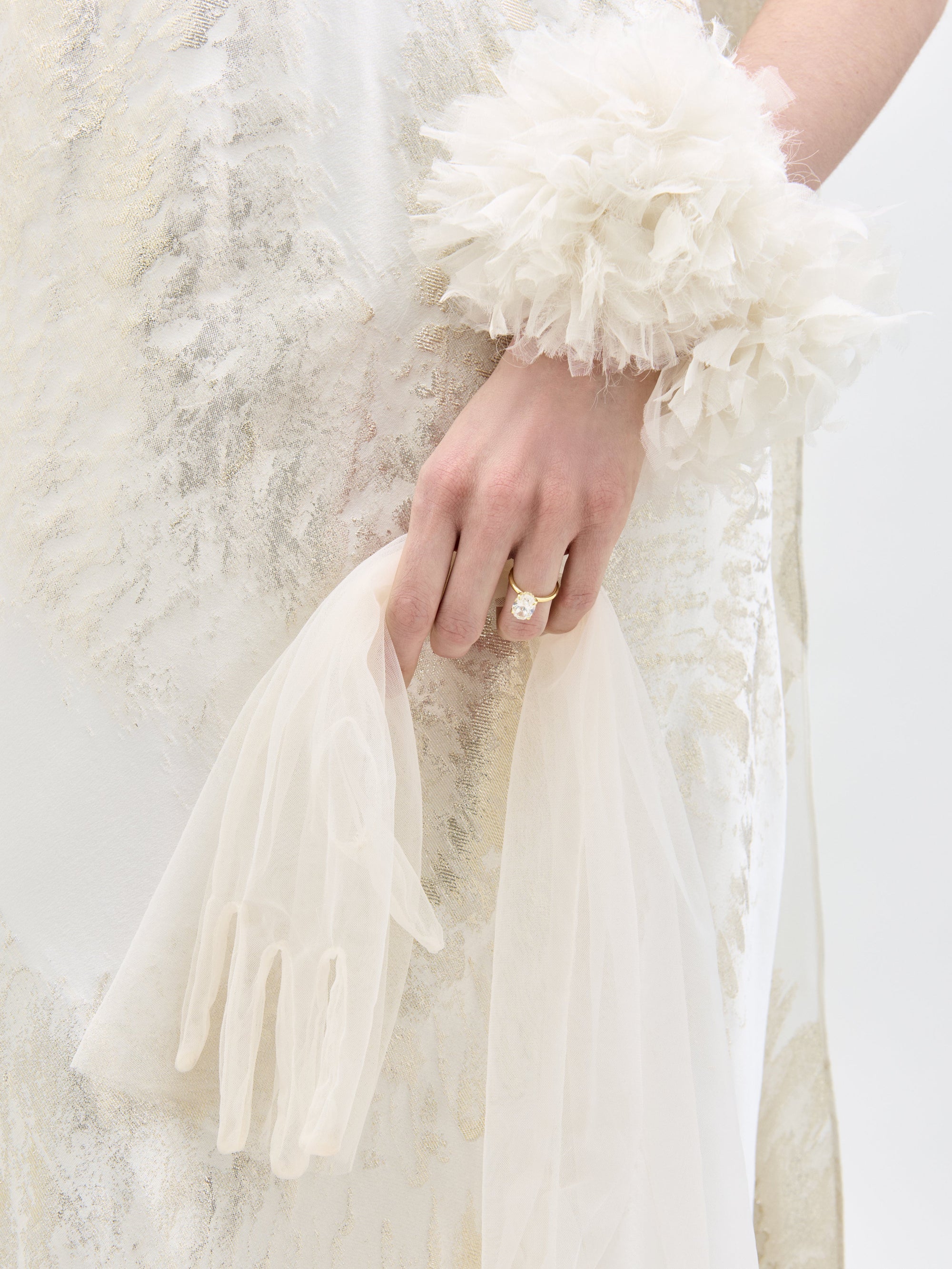 A woman poses in the Sabina Dress, a white and beige flowing gown featuring a high neckline and cape-like sleeves, set against a plain backdrop.