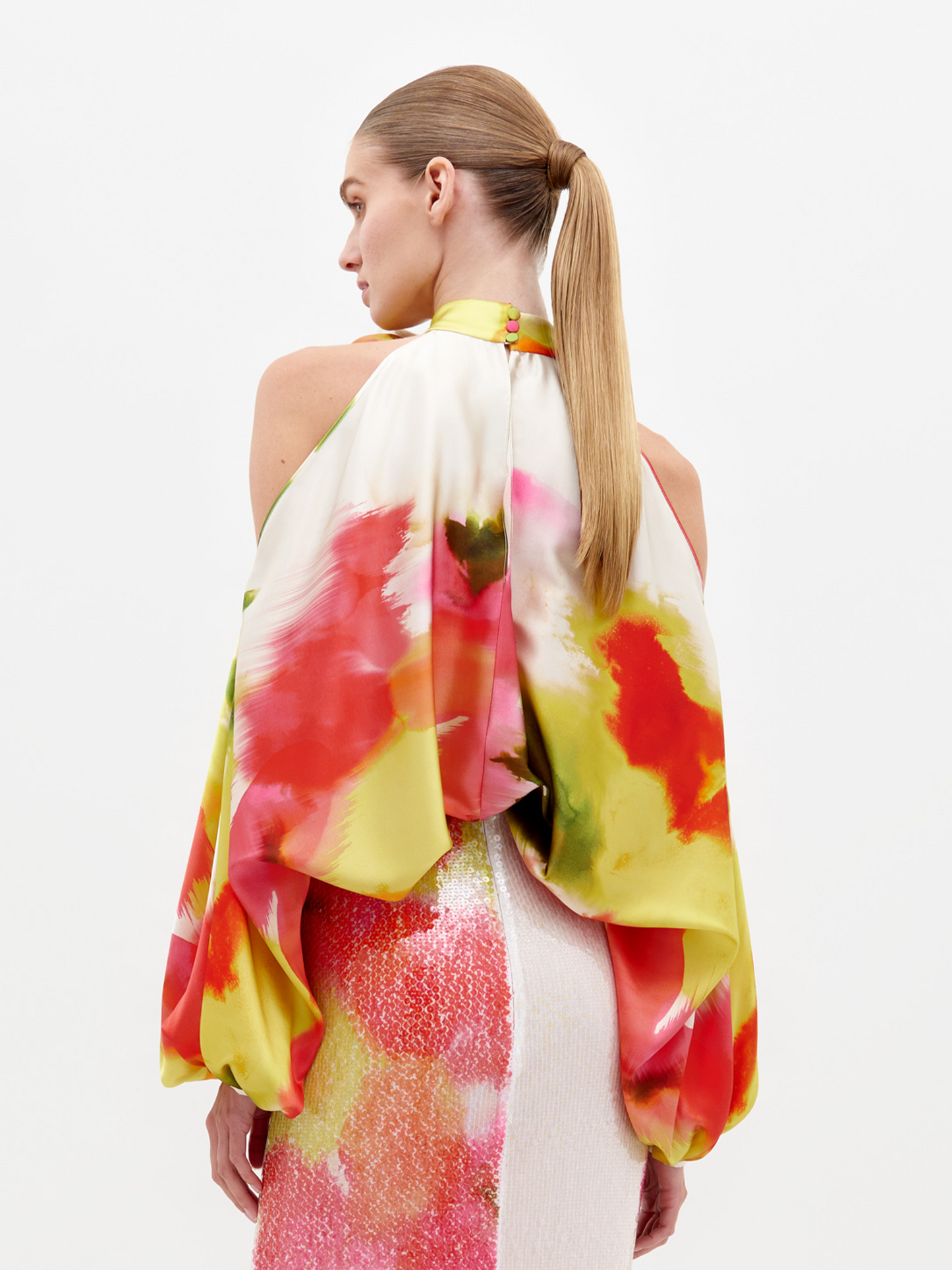 A woman models the Safira Blouse, featuring a colorful watercolor floral pattern, halter neckline, and a stylish floral appliqué on the shoulder, set against a plain white background.