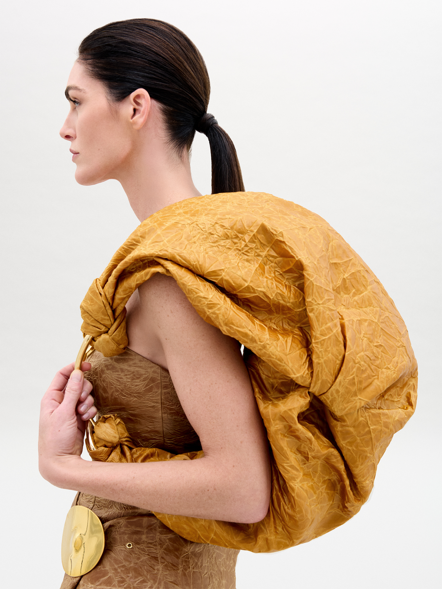 The Sofia Bag Maxi is a brown bag with a crinkled texture, made from Italian fabric. It features a gold circular handle and knotted details on both sides, shown against a plain white background.