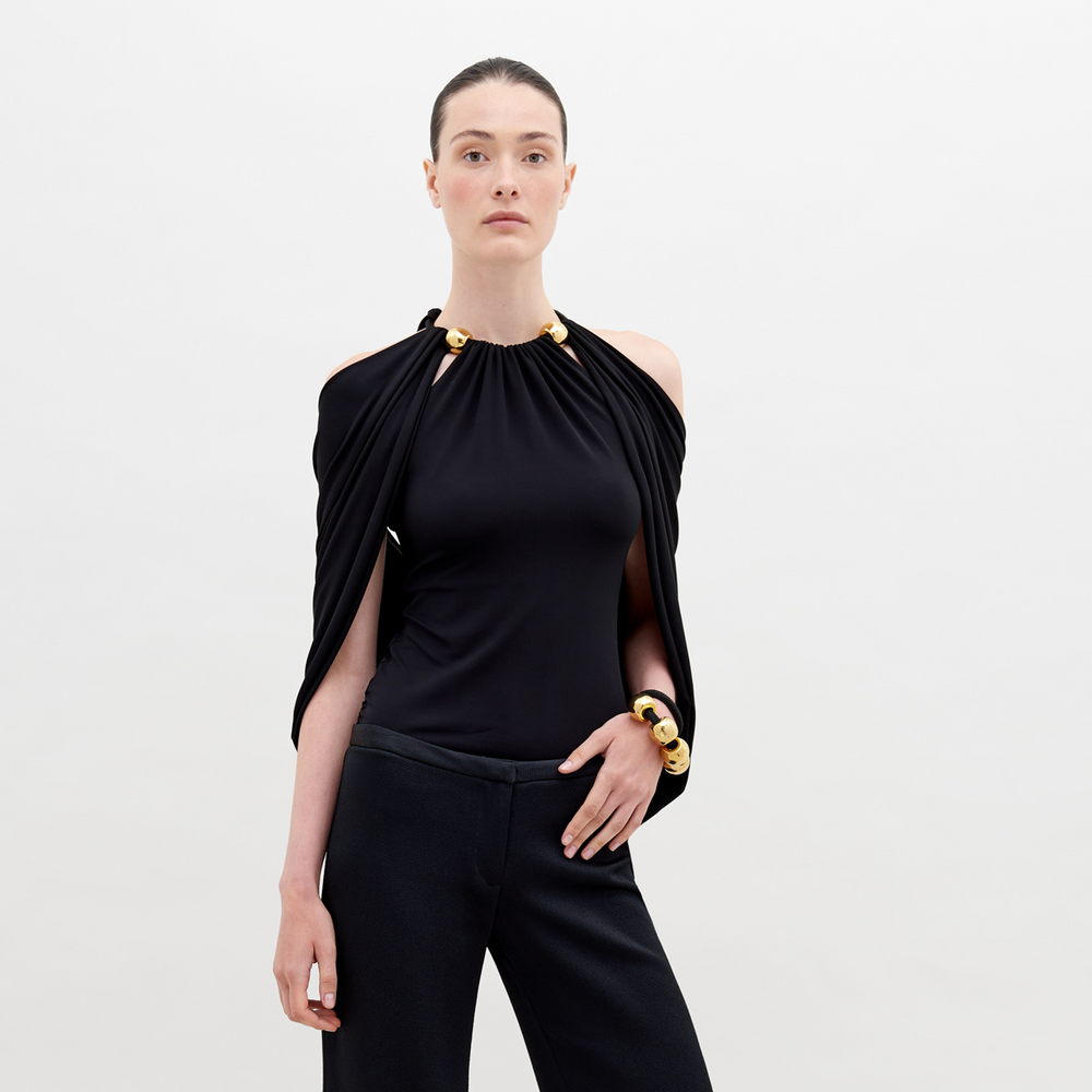 A person with tied-back hair wears a Tabatha Blouse Black with gold accents and a matching gold bracelet, standing against a plain white background. This stylish ensemble is available for pre-order now, with a ship by date of November 15th, 2024.