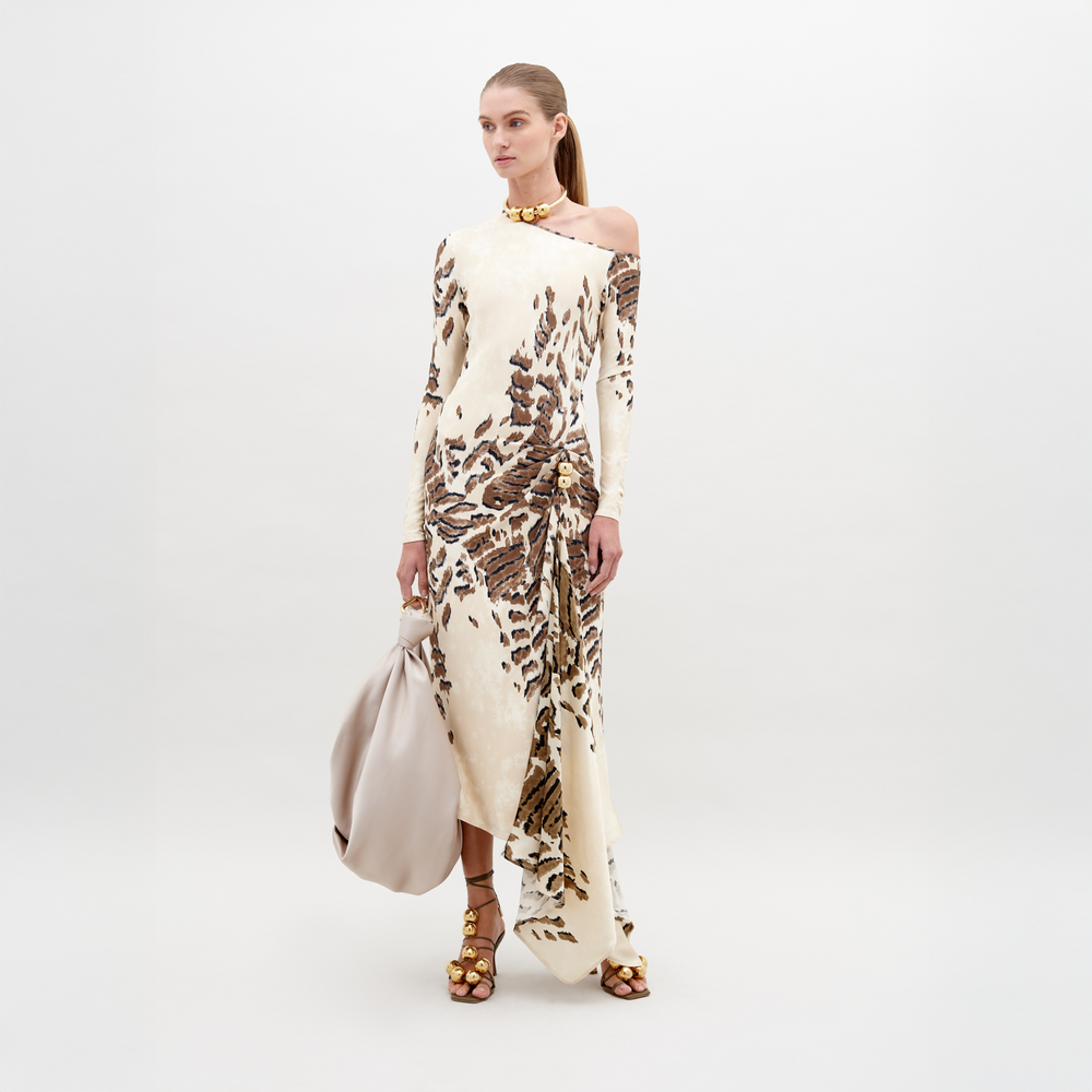 A woman in an off-shoulder, floor-length Tahia Dress Beige Abstract Animal Print stands against a plain background, holding a large beige bag in one hand. She is wearing gold sandals and a gold choker. Mark your calendars for November 15th—this stunning look is available for pre-order ahead of its 2024 launch.
