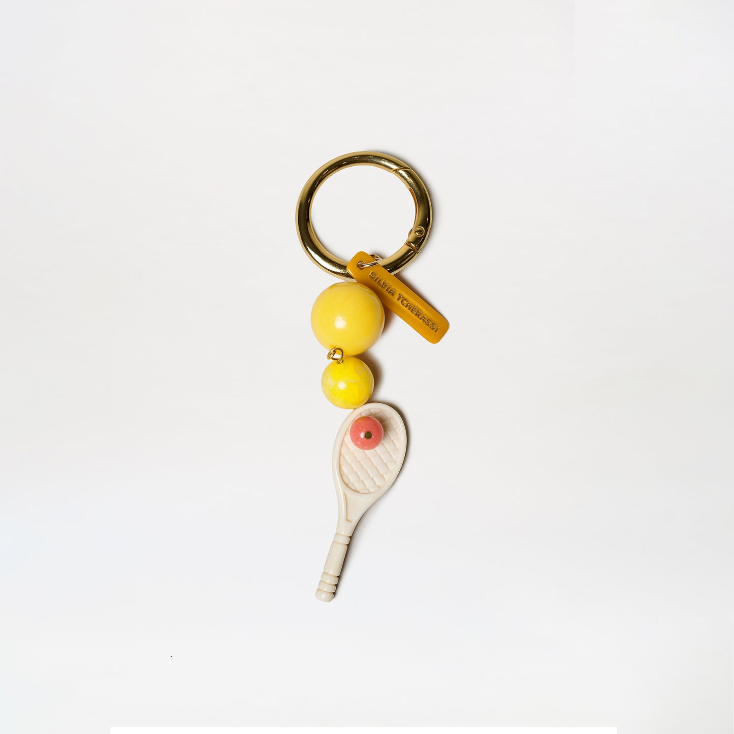The Deuce Charm is a keychain featuring a gold ring, two yellow beads, an orange LOEWE FOUNDATION tag, and a miniature white tennis racket charm with an orange dot at its center.