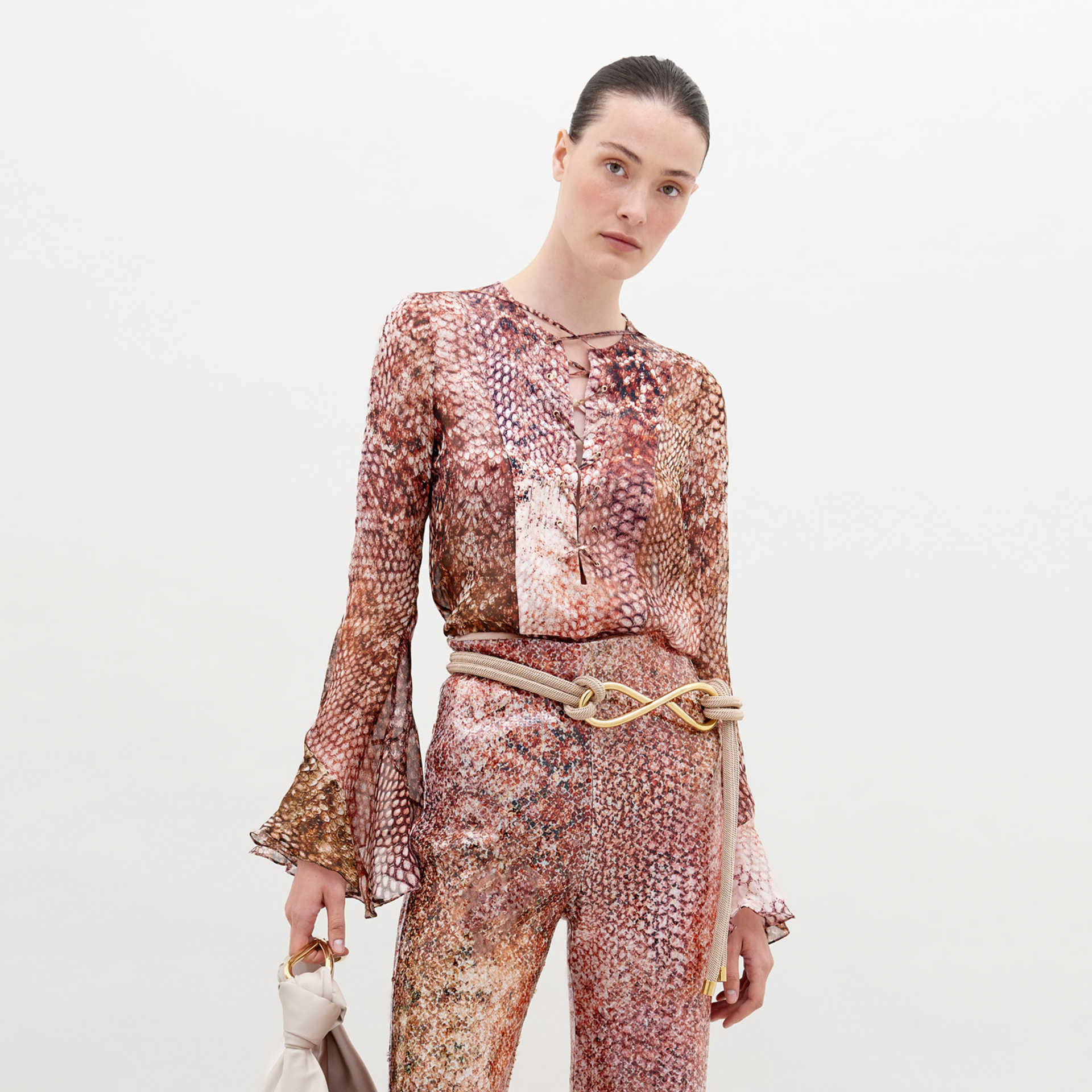 A person is wearing a Thea Blouse Peach Animal Print with flared cuffs and matching pants. They have a neutral expression and are holding a white accessory, available for pre-order now. Secure your piece before November 15th, 2024.