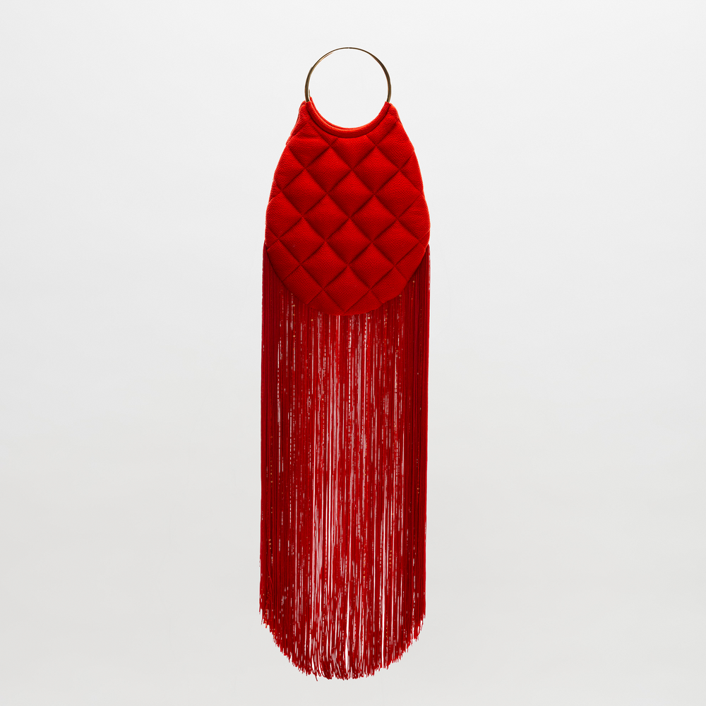 The Toblach Bag is a red quilted handbag made from Italian fabric, featuring metallic top handles and long red fringe along the bottom, displayed against a plain white background.