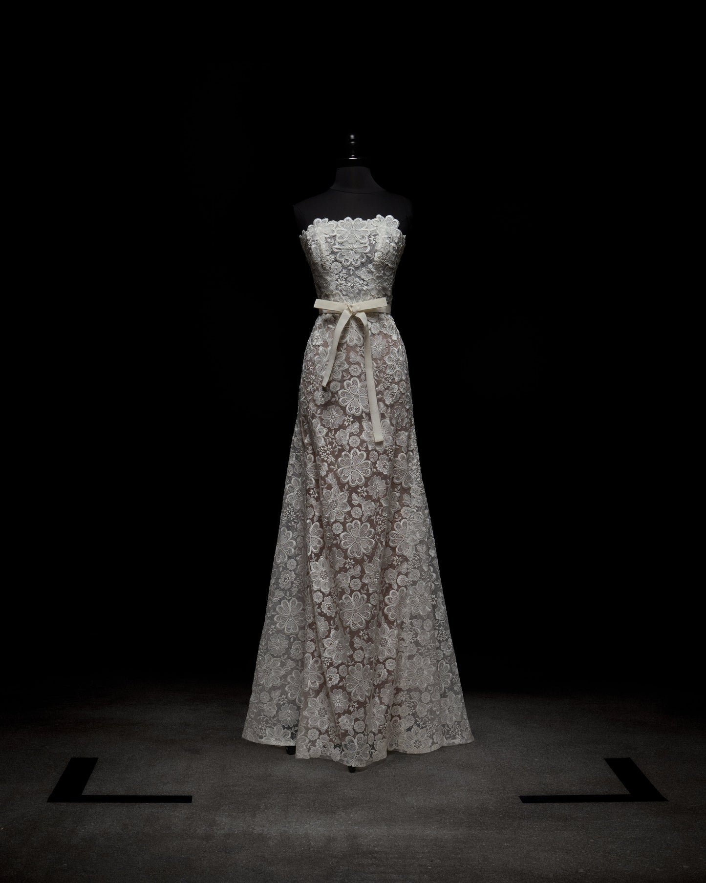 A woman poses against a plain white background in the Valencia Dress, which features a strapless design with lace floral patterns. She complements it with sheer gloves.