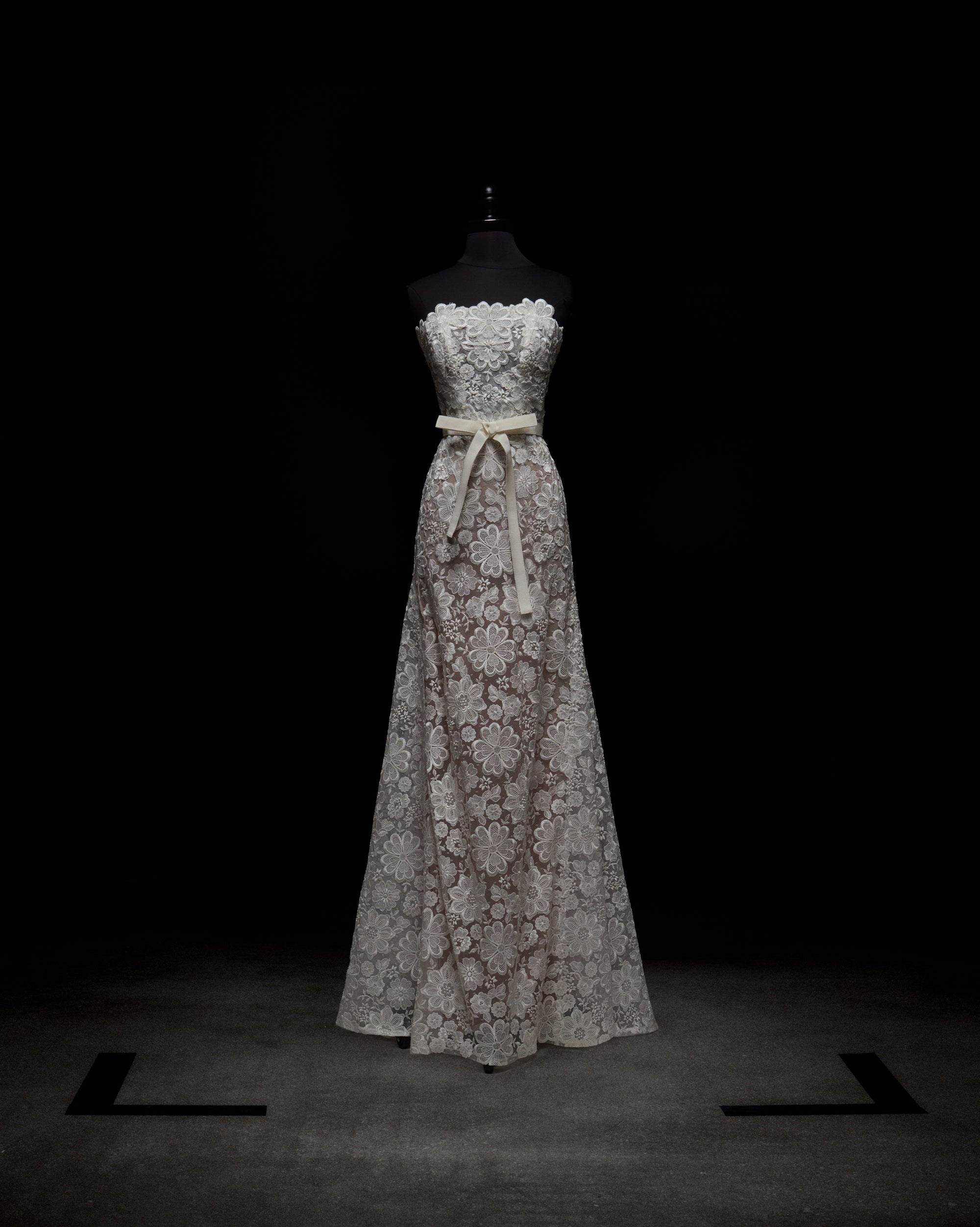 A woman poses against a plain white background in the Valencia Dress, which features a strapless design with lace floral patterns. She complements it with sheer gloves.