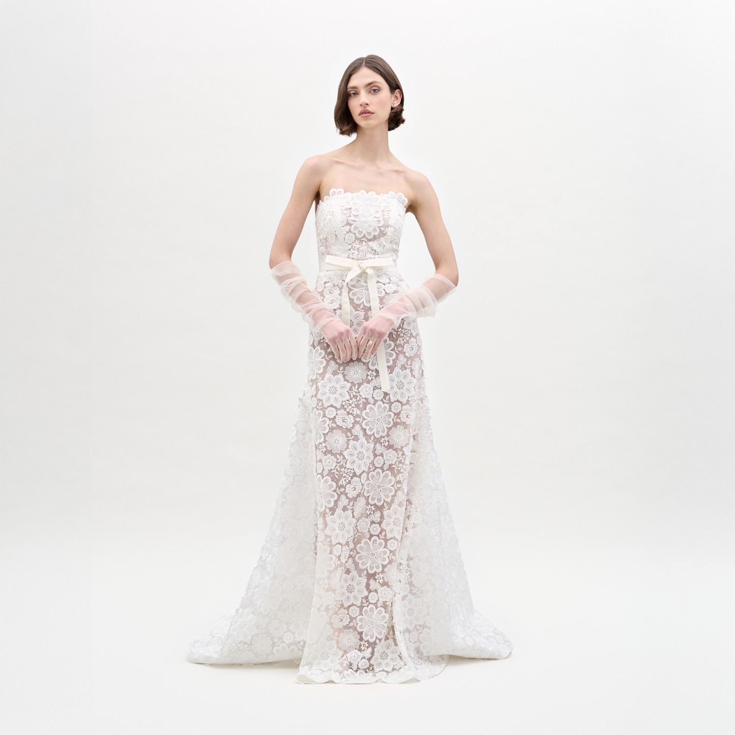 A woman poses against a plain white background in the Valencia Dress, which features a strapless design with lace floral patterns. She complements it with sheer gloves.