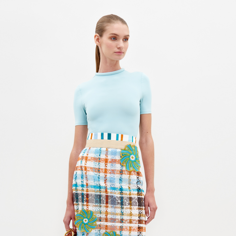 A woman dressed in a fitted Varinia Tshirt Turquoise and a multicolored plaid skirt stands against a plain white background, holding a small brown item in her right hand.