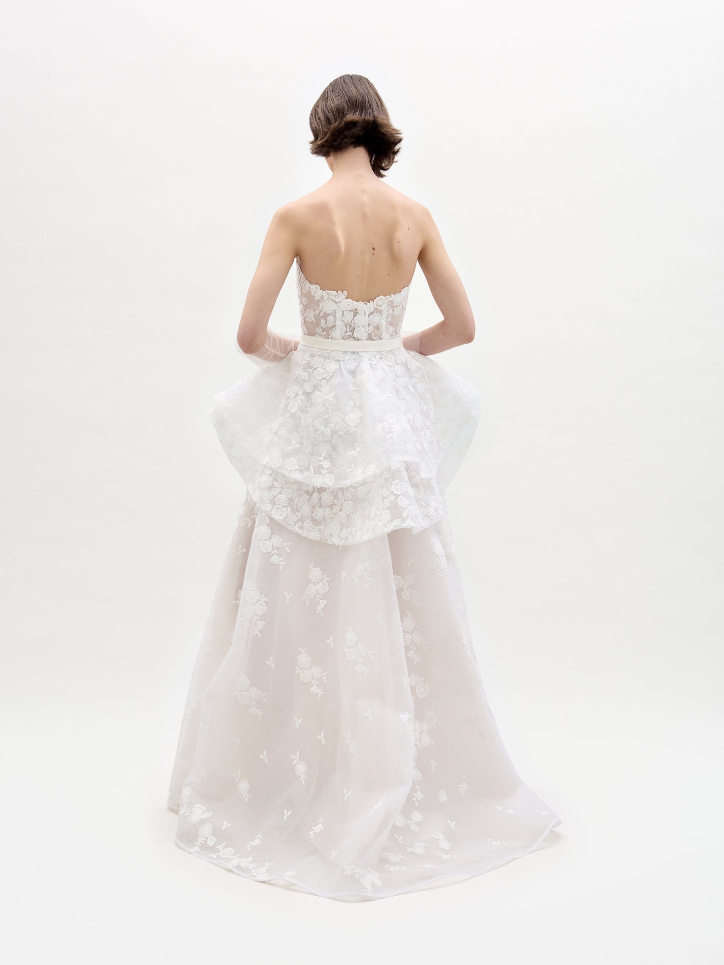 A woman in the Viviana Dress, a strapless white wedding gown featuring floral lace details and a peplum design, stands against a light plain background.