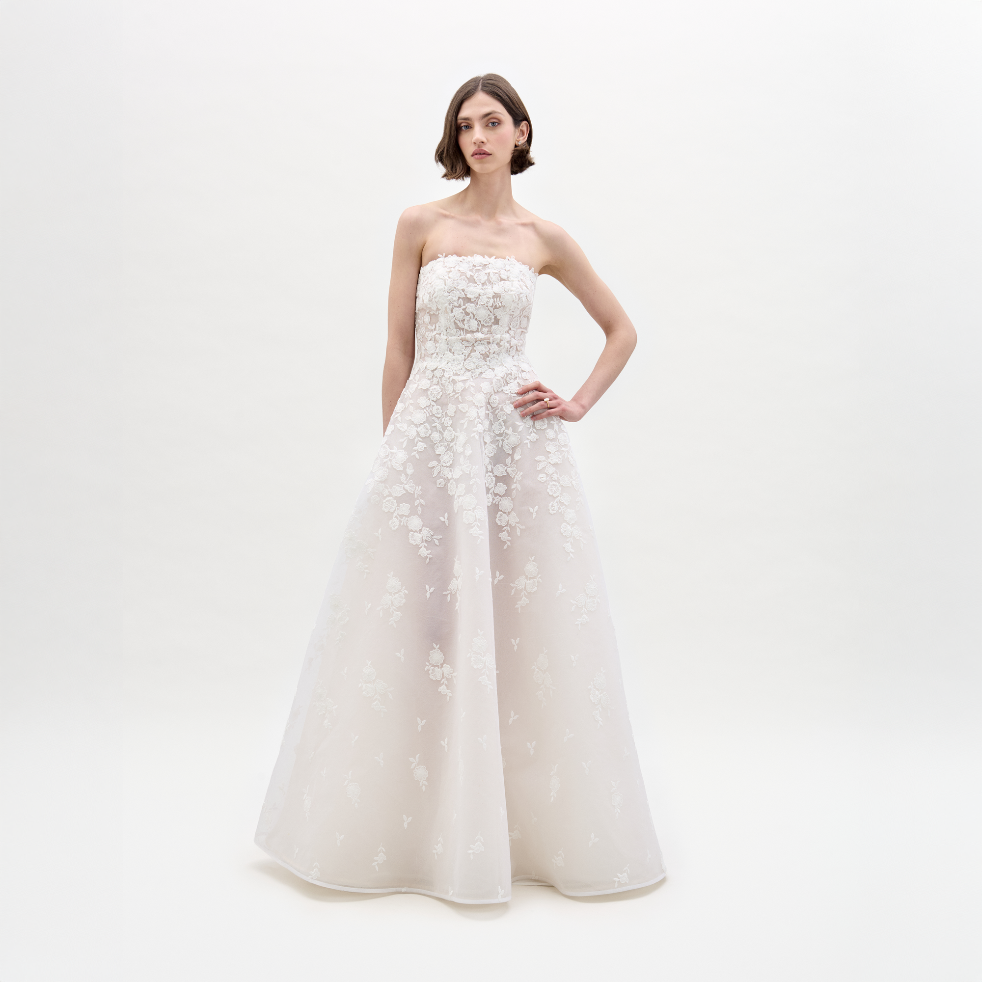 A woman in the Viviana Dress, a strapless white wedding gown featuring floral lace details and a peplum design, stands against a light plain background.