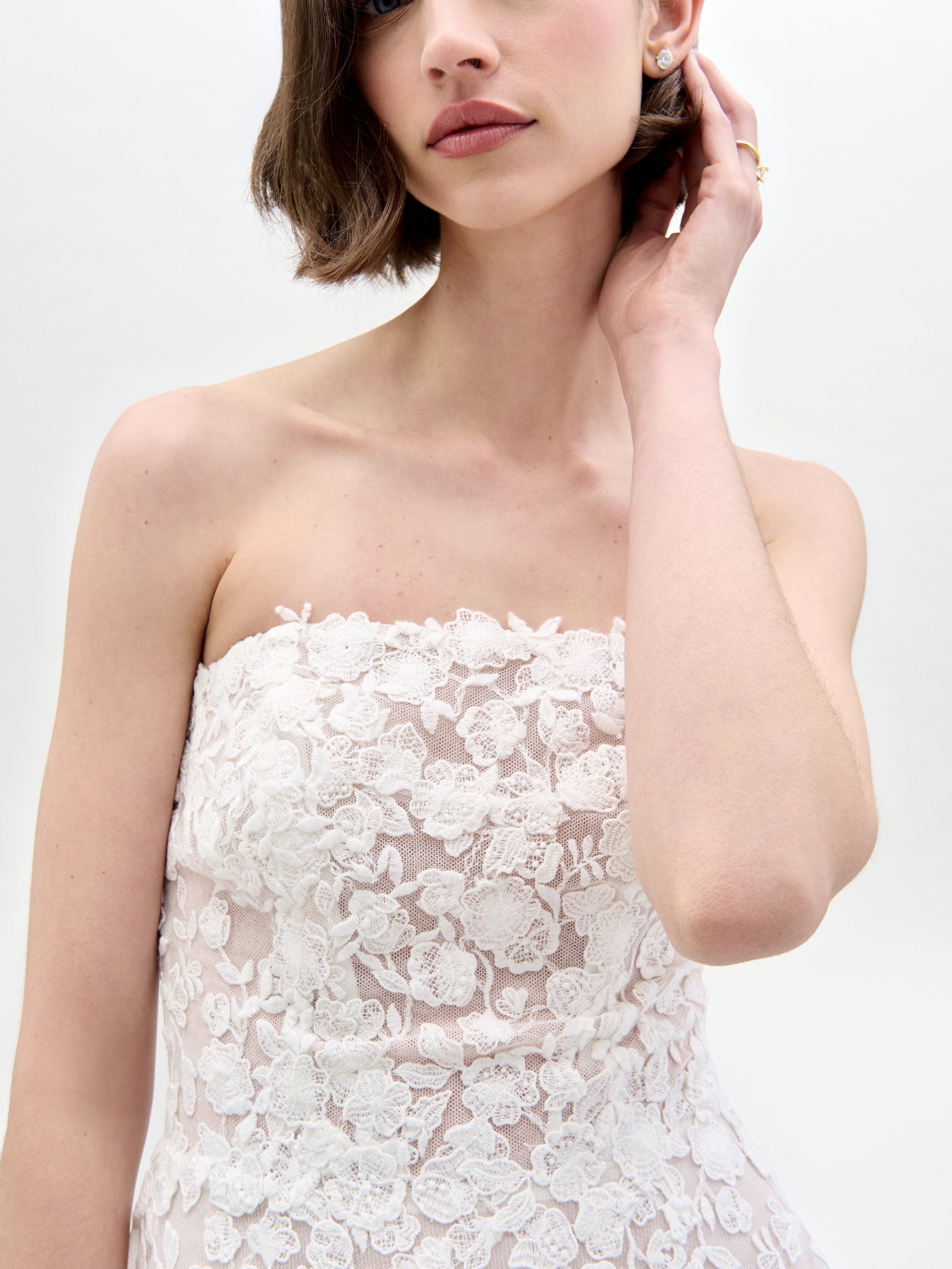 A woman in the Viviana Dress, a strapless white wedding gown featuring floral lace details and a peplum design, stands against a light plain background.