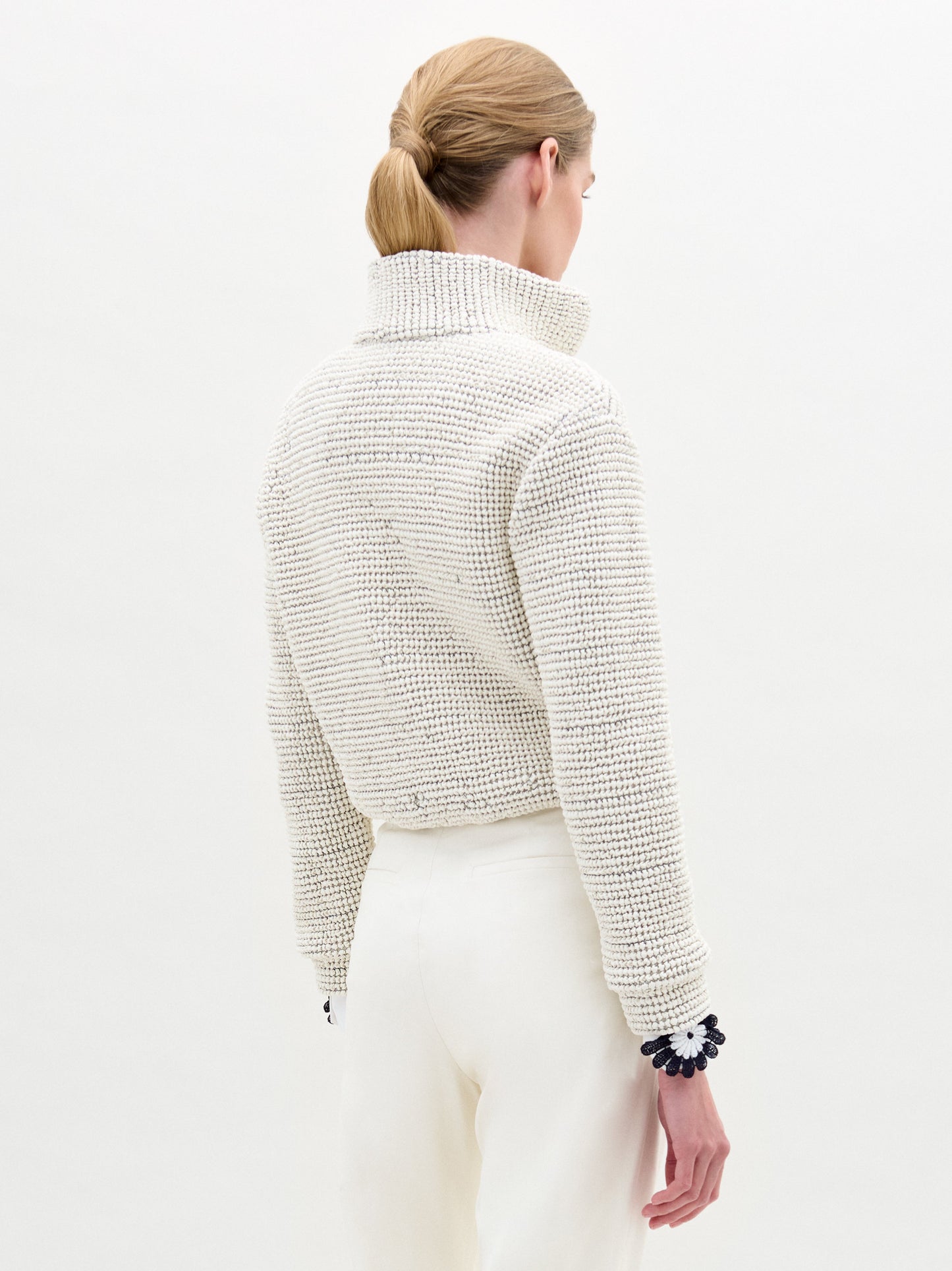 A woman stands against a plain white background wearing the Zalta Jacket—a white textured eco leather zip-up—over a white collared shirt and high-waisted white trousers.