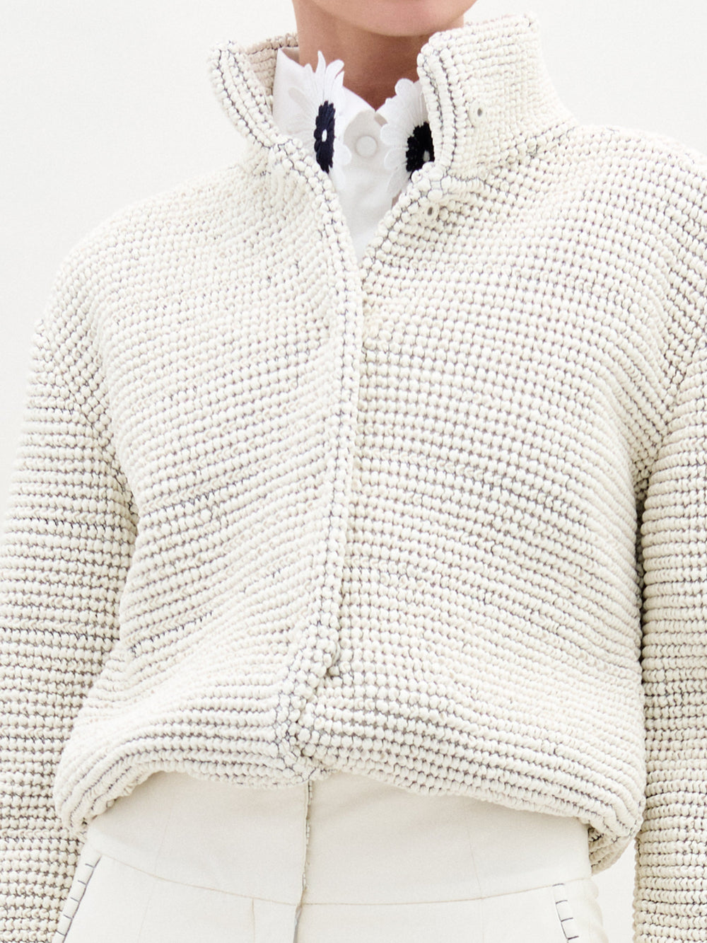 A woman stands against a plain white background wearing the Zalta Jacket—a white textured eco leather zip-up—over a white collared shirt and high-waisted white trousers.