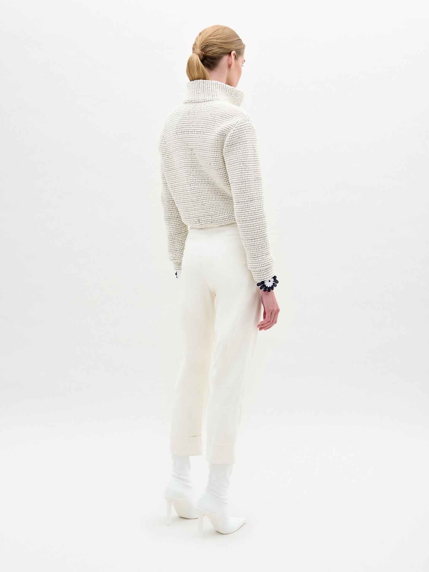 A person stands against a plain light background, wearing a textured white sweater, white collared shirt, the Moad Pant—an Italian fabric high-waisted cropped pant—and white heeled boots.