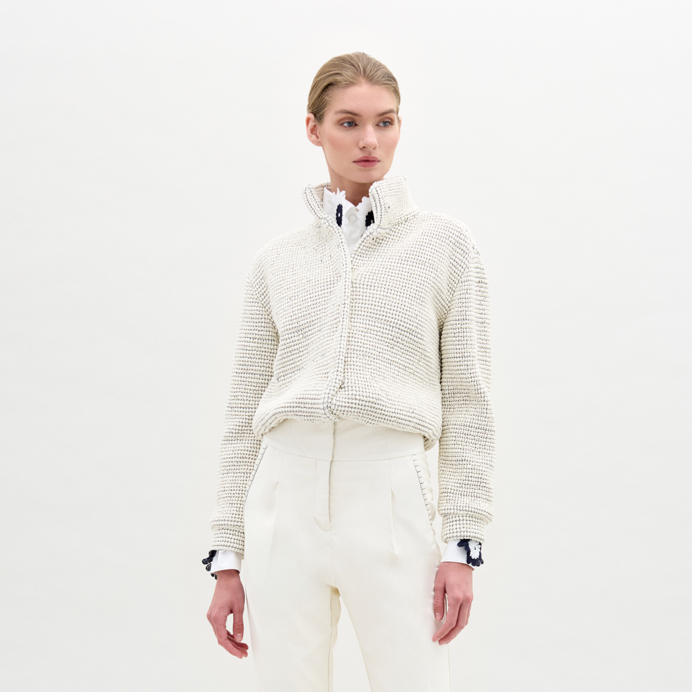 A woman stands against a plain white background wearing the Zalta Jacket—a white textured eco leather zip-up—over a white collared shirt and high-waisted white trousers.