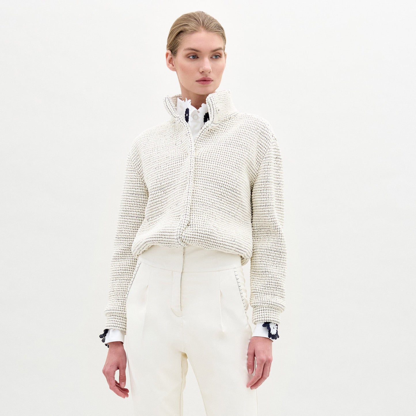 A woman wears the Zalta Jacket, a textured white cropped style, over a white collared shirt and high-waisted trousers, standing against a plain light background.