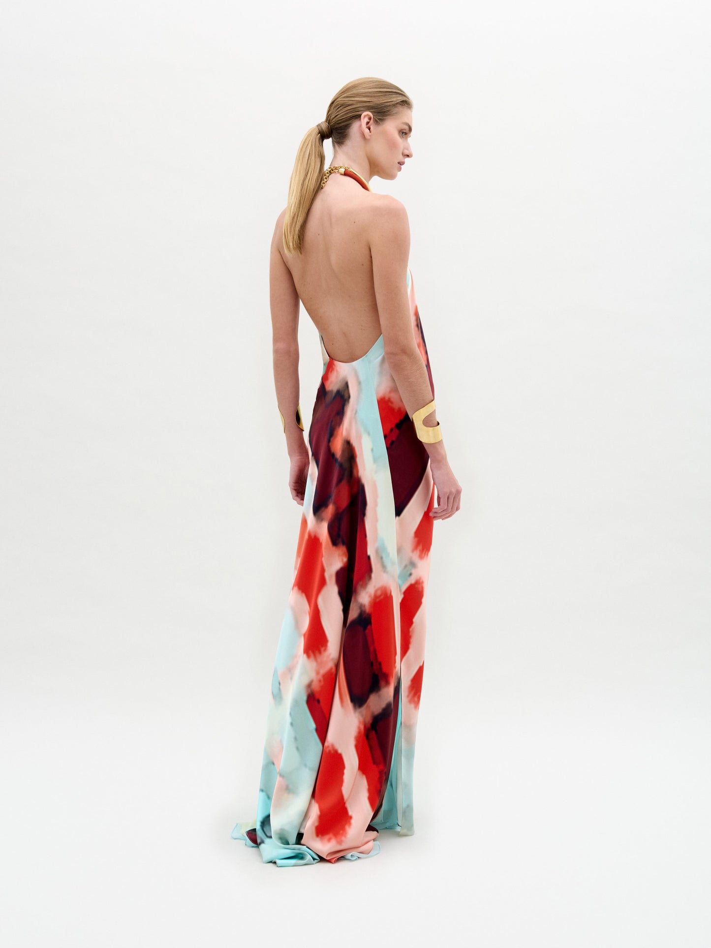A woman poses against a white background wearing the Zeffa Dress, a sleeveless, floor-length silk maxi with an abstract red, aqua, and white pattern, paired with gold jewelry.