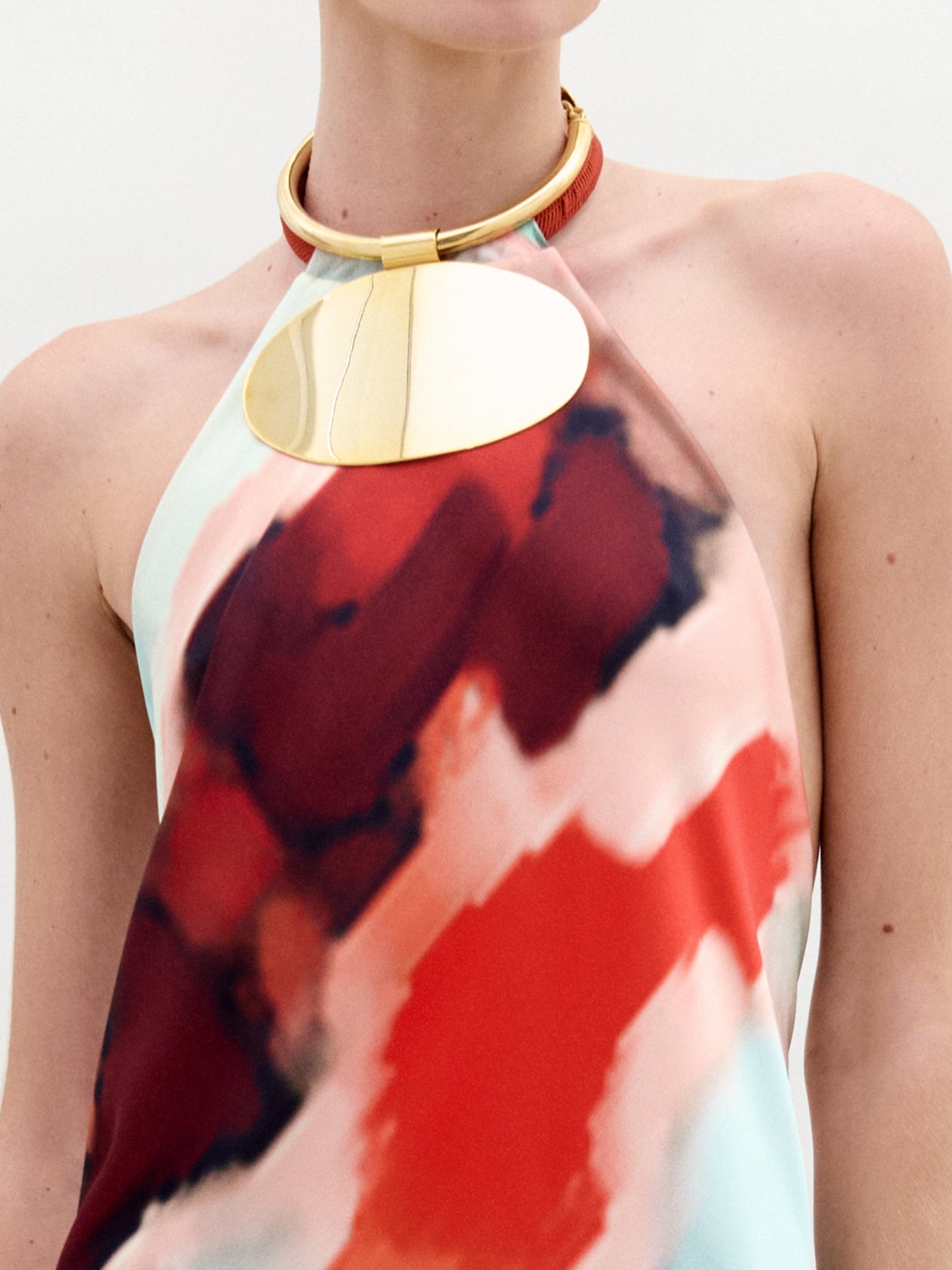 A woman poses against a white background wearing the Zeffa Dress, a sleeveless, floor-length silk maxi with an abstract red, aqua, and white pattern, paired with gold jewelry.