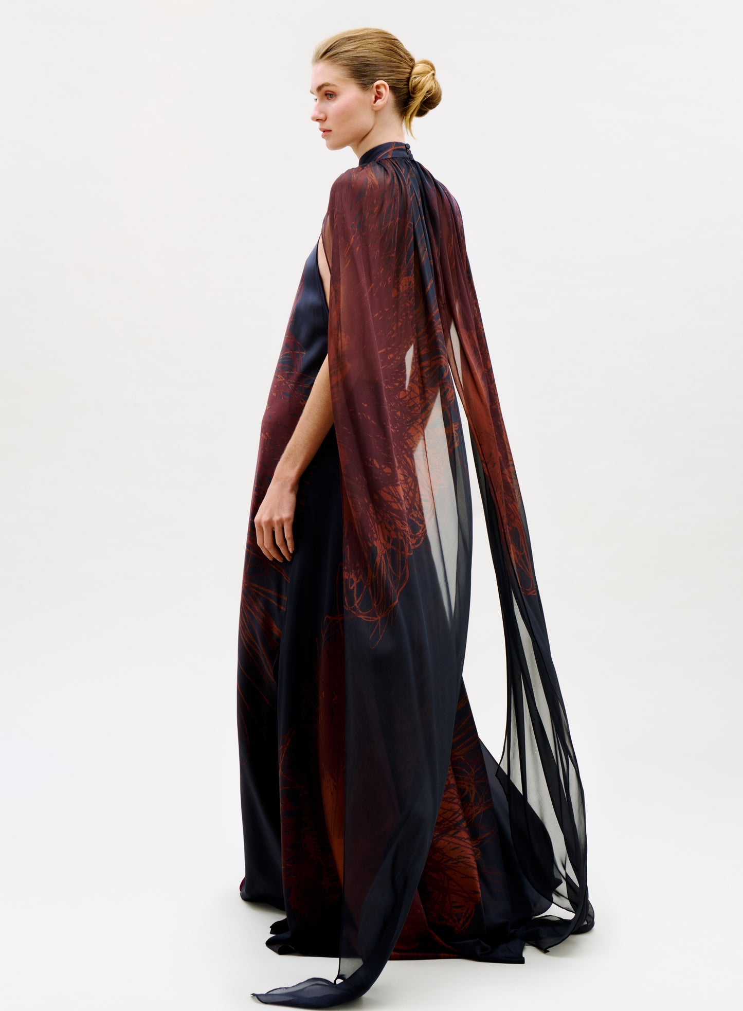 A woman with blonde hair in a low bun models the Sabina Dress, a flowing sheer black and dark red gown with a dramatic cape, standing in profile against a plain white background.