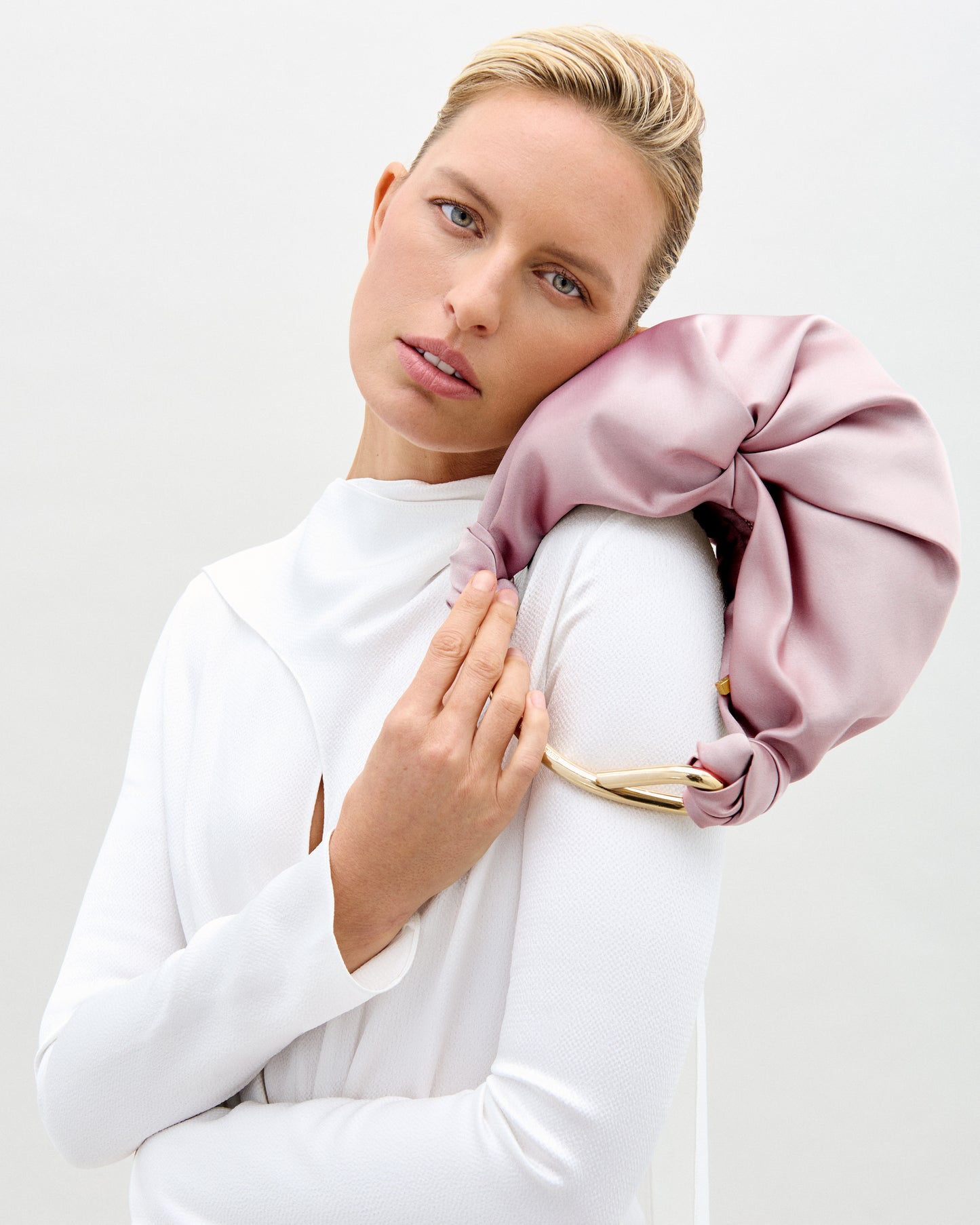 A person with short blonde hair in a white outfit gently leans their head on the Sofia Bag (Small), a large, pale pink satin accessory with gold detail and handle, crafted from luxurious Italian fabric, set against a light background.