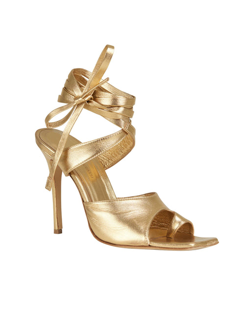 A pair of Golda Heels Gold with a bow on the ankle, perfect for adding an extra touch of elegance to any outfit.