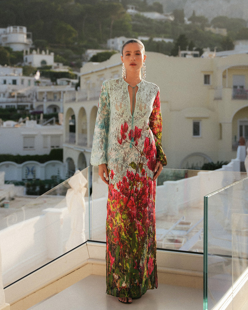 Nane Miller stands on a balcony in a vibrant floral dress against a backdrop of Mediterranean-style buildings and lush hills, conveying elegance and tranquility.