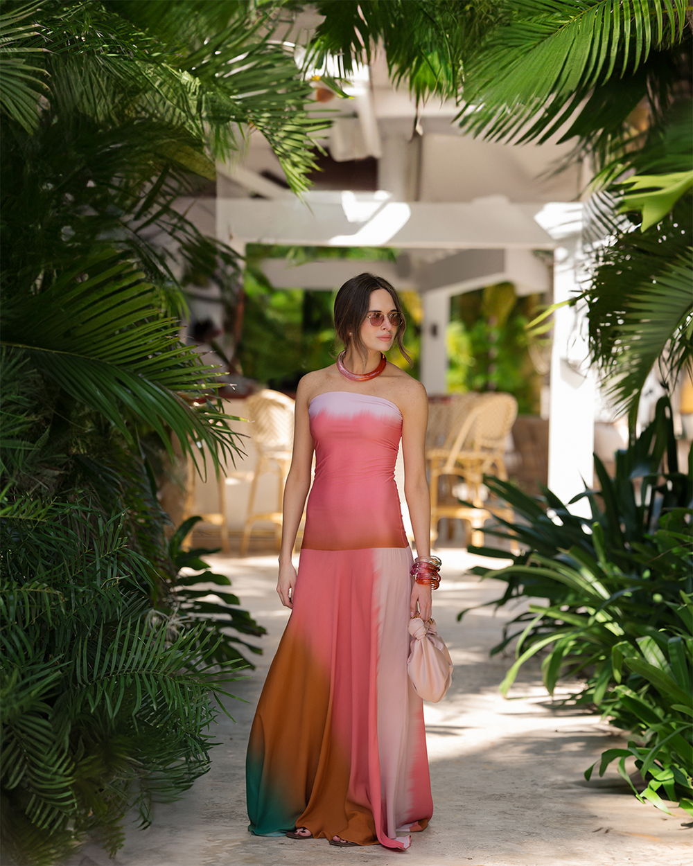 Sofia Tcherassi in a colorful strapless dress walks through a lush, tropical setting. She's carrying a purse, wearing sunglasses, and appears relaxed.