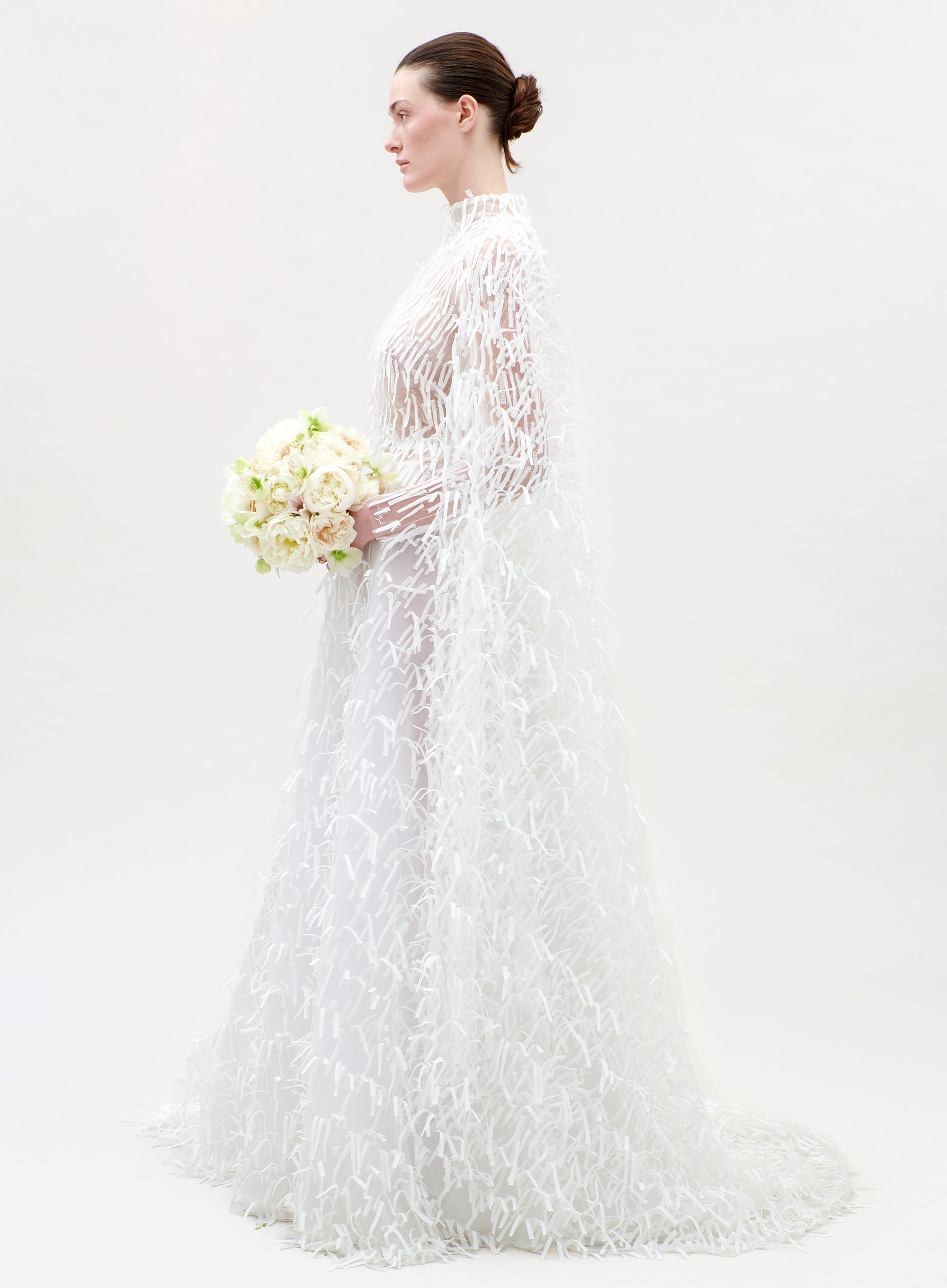 Woman in a white lace wedding dress holding a bouquet on a plain background
