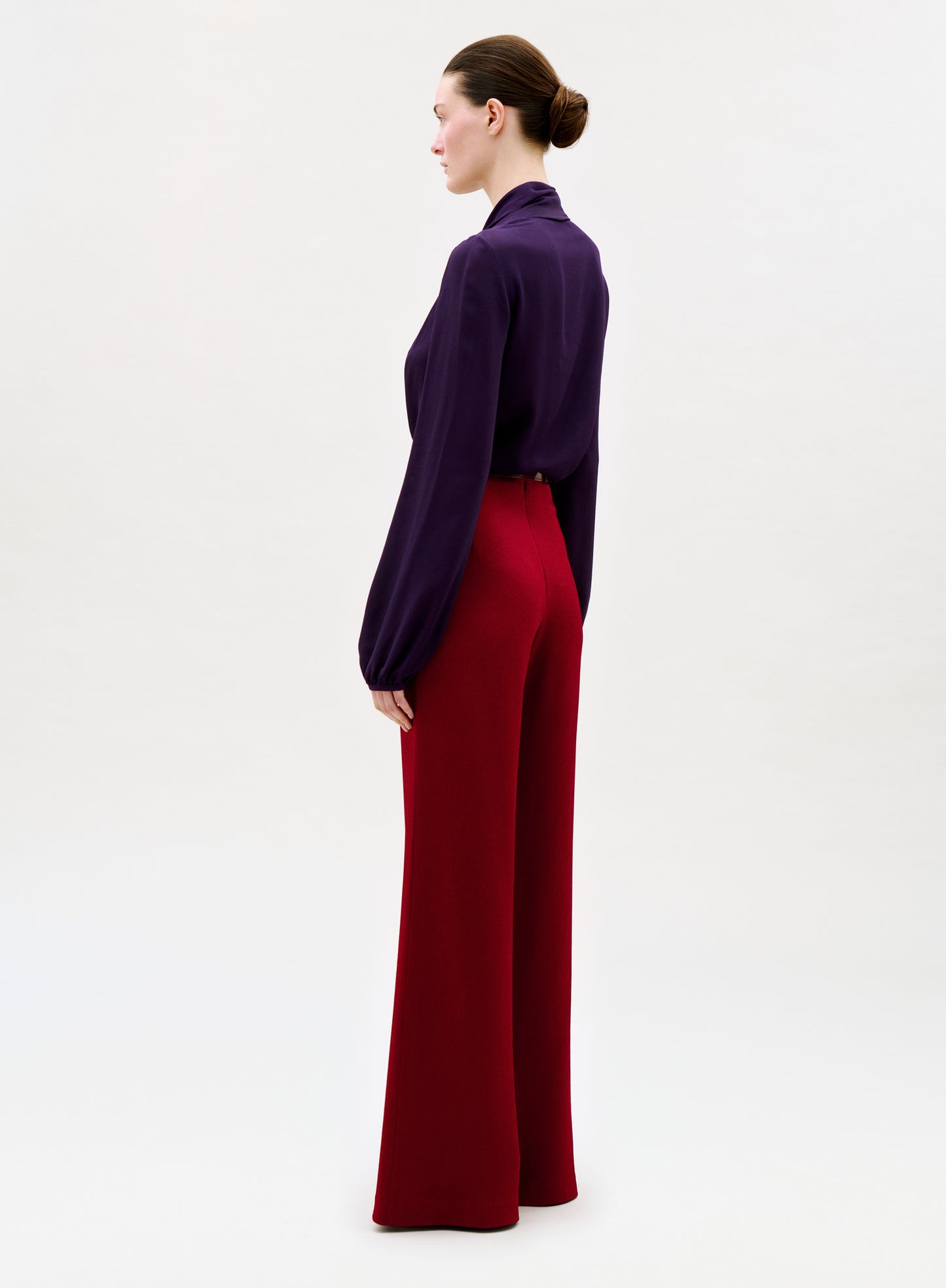 A woman in profile stands against a plain light background, wearing a dark purple long-sleeved blouse and high-waisted Palermo Pant in wide-leg red. Her hair is styled neatly in a bun.