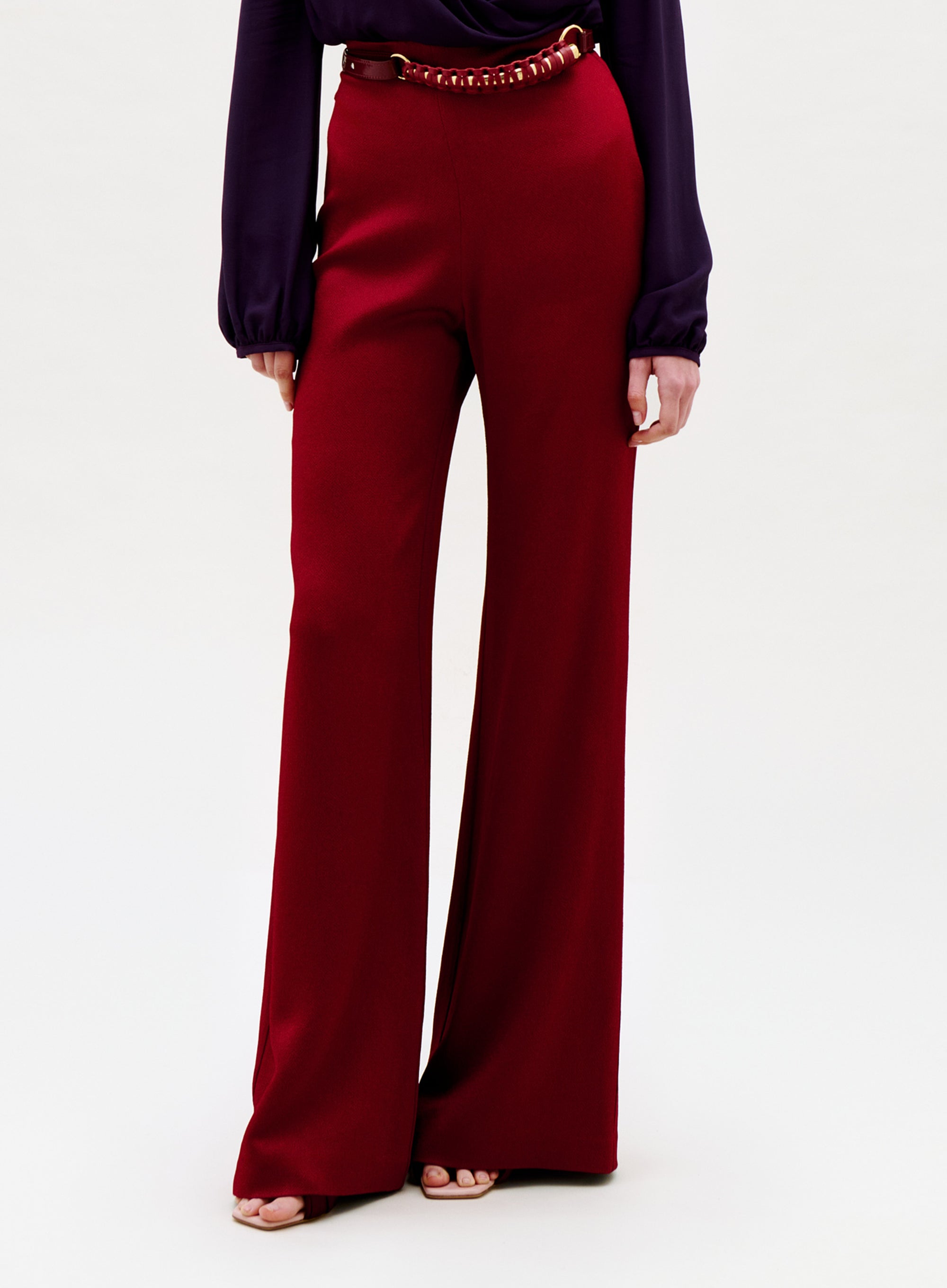 Someone wearing Palermo Pant—high-waisted, wide-leg red pants with a thin belt—paired with a long-sleeved dark purple top, stands against a plain light background.