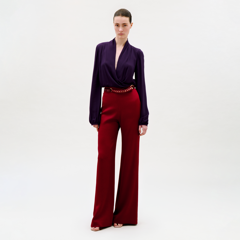 A woman stands against a plain light background, wearing a deep purple long-sleeve blouse with a plunging neckline and high-waisted, wide-leg Palermo Pant in red, accessorized with a red belt and open-toed shoes.
