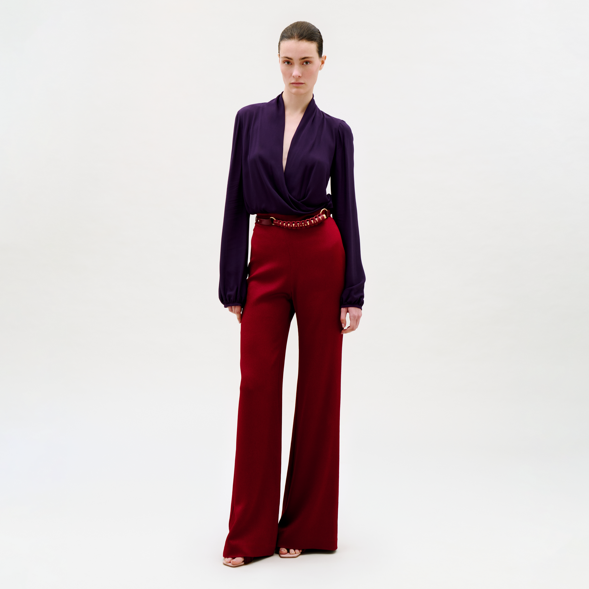A woman stands against a plain light background, wearing a deep purple long-sleeve blouse with a plunging neckline and high-waisted, wide-leg Palermo Pant in red, accessorized with a red belt and open-toed shoes.