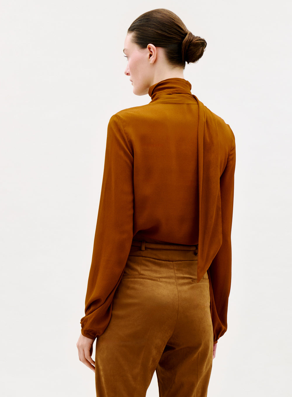 A person with brown hair in a neat bun is shown from the back, wearing the Alanis Blouse—a long-sleeved, high-neck brown blouse with a draped scarf detail—paired with matching brown pants against a plain light background.
