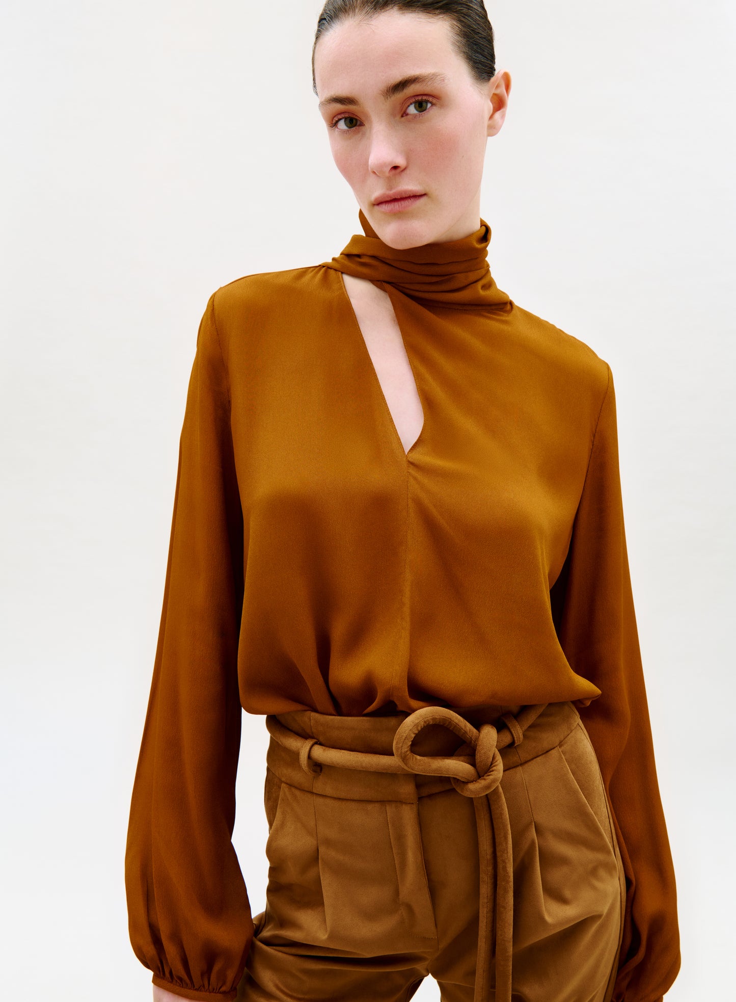 A woman wearing the Alanis Blouse, a silky rust-colored top with a high neck and asymmetrical cut-out, paired with high-waisted, belted tan trousers, stands against a plain white background.
