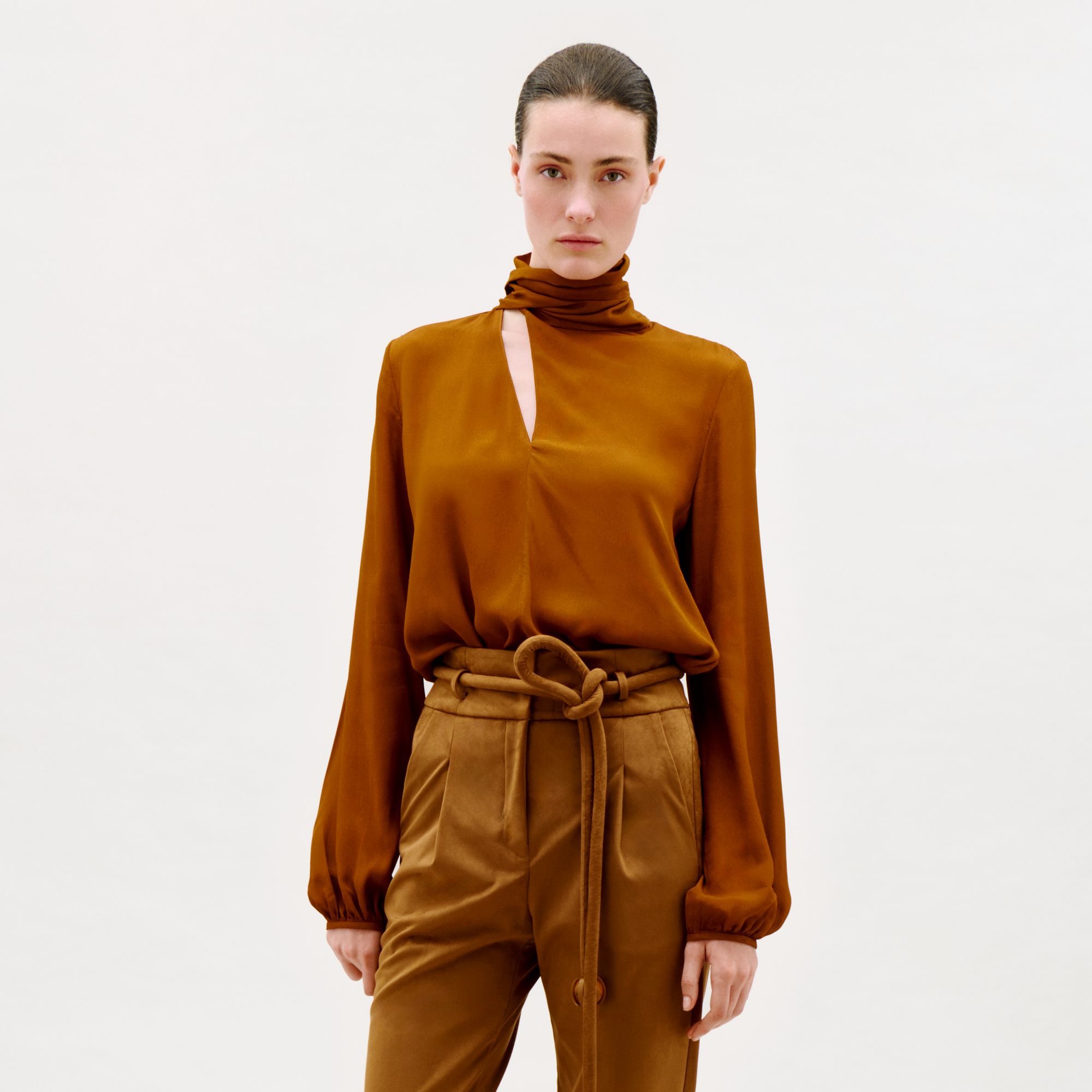 A person stands against a plain light background, wearing the Alanis Blouse—a copper brown long-sleeve top with a high neck and front slit—paired with matching high-waisted pants tied with a fabric belt.