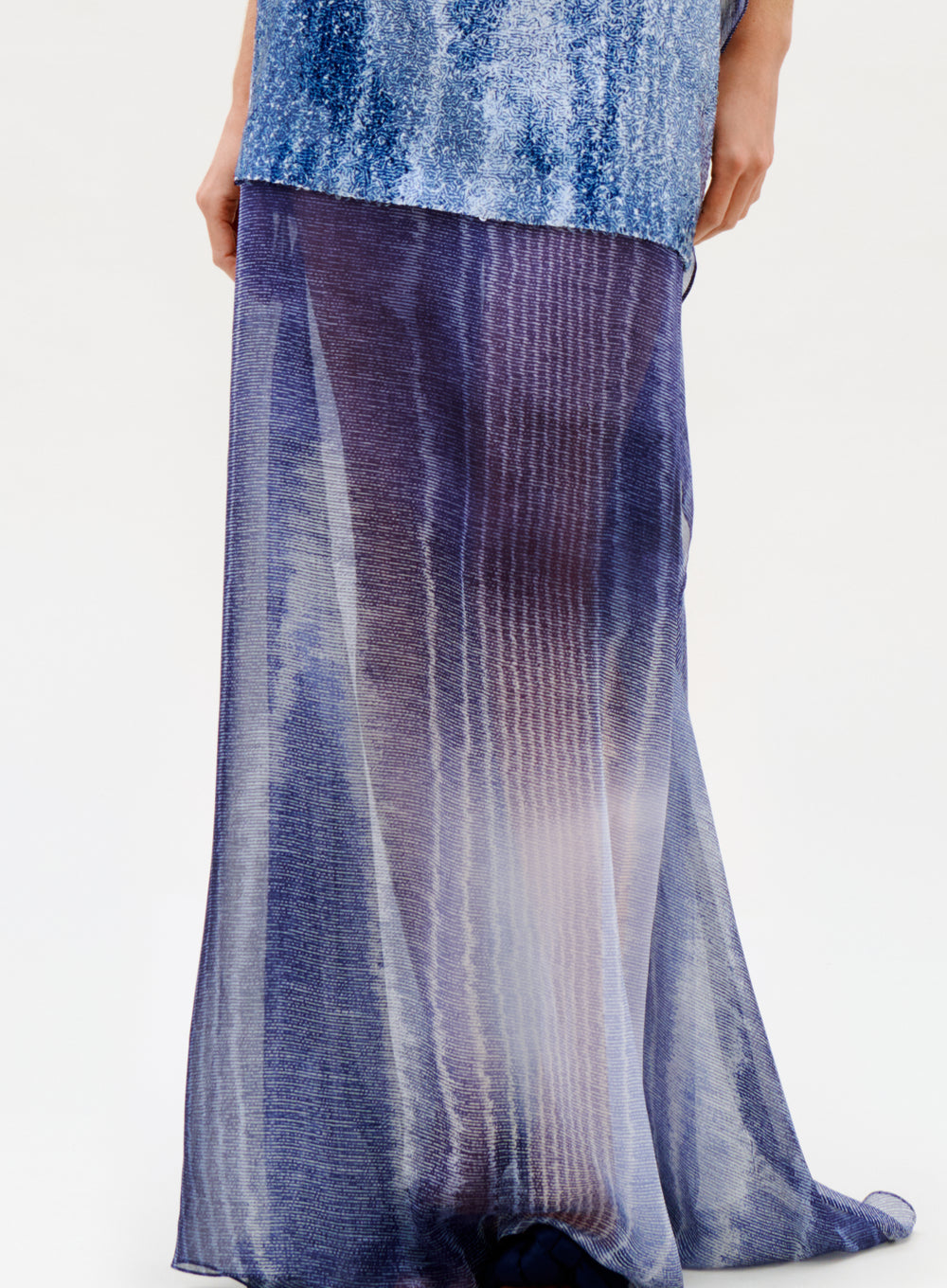 Wearing the Amalia Skirt—a long, flowing silk maxi with a sheer blue and purple gradient and subtle vertical stripes—paired with a shimmery blue top, this person stands against a plain light background.