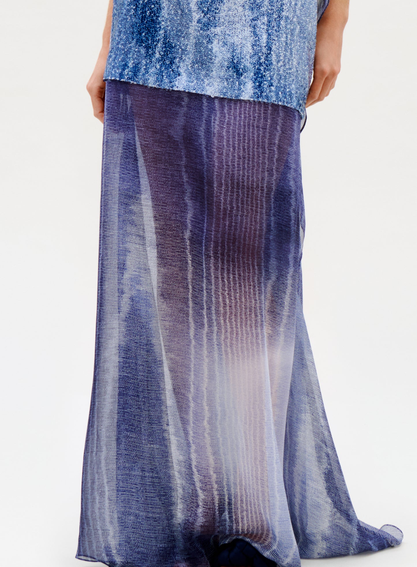 Wearing the Amalia Skirt—a long, flowing silk maxi with a sheer blue and purple gradient and subtle vertical stripes—paired with a shimmery blue top, this person stands against a plain light background.