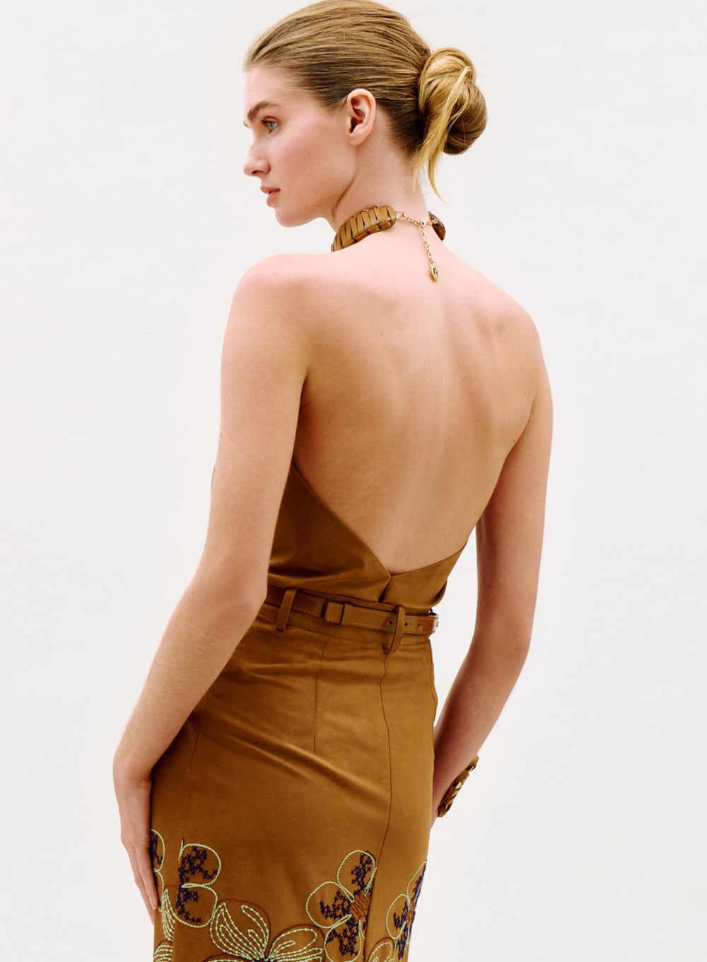A woman with blonde hair in a low bun wears the Ann Blouse, featuring an open back and embroidered floral details, standing against a plain light background.