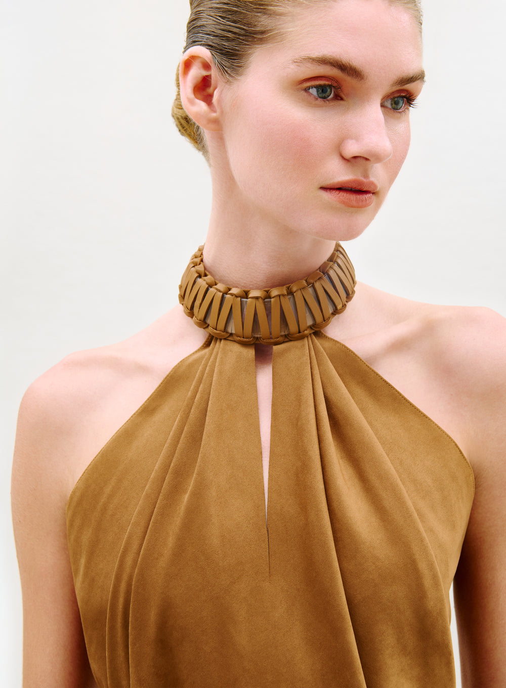 A woman with light skin and blue eyes, her hair slicked back neatly, wears the Ann Blouse—a tan halter style with a high, wide textured collar—while looking slightly to the side against a plain light background.