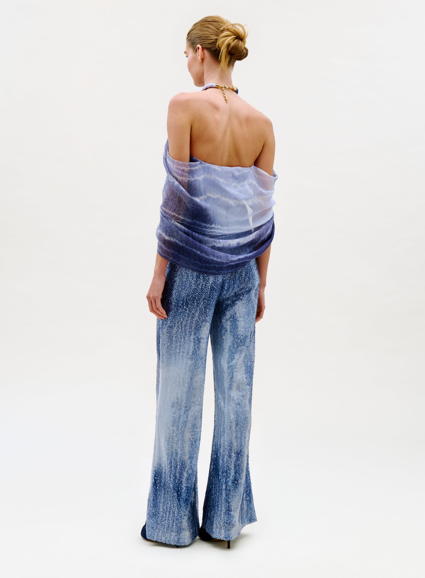A woman stands facing away, styled in a sheer off-shoulder blue top paired with the high-waisted, wide-leg Avellino Pant. Her hair is neatly tied in a bun and she wears a necklace, set against a plain light background.