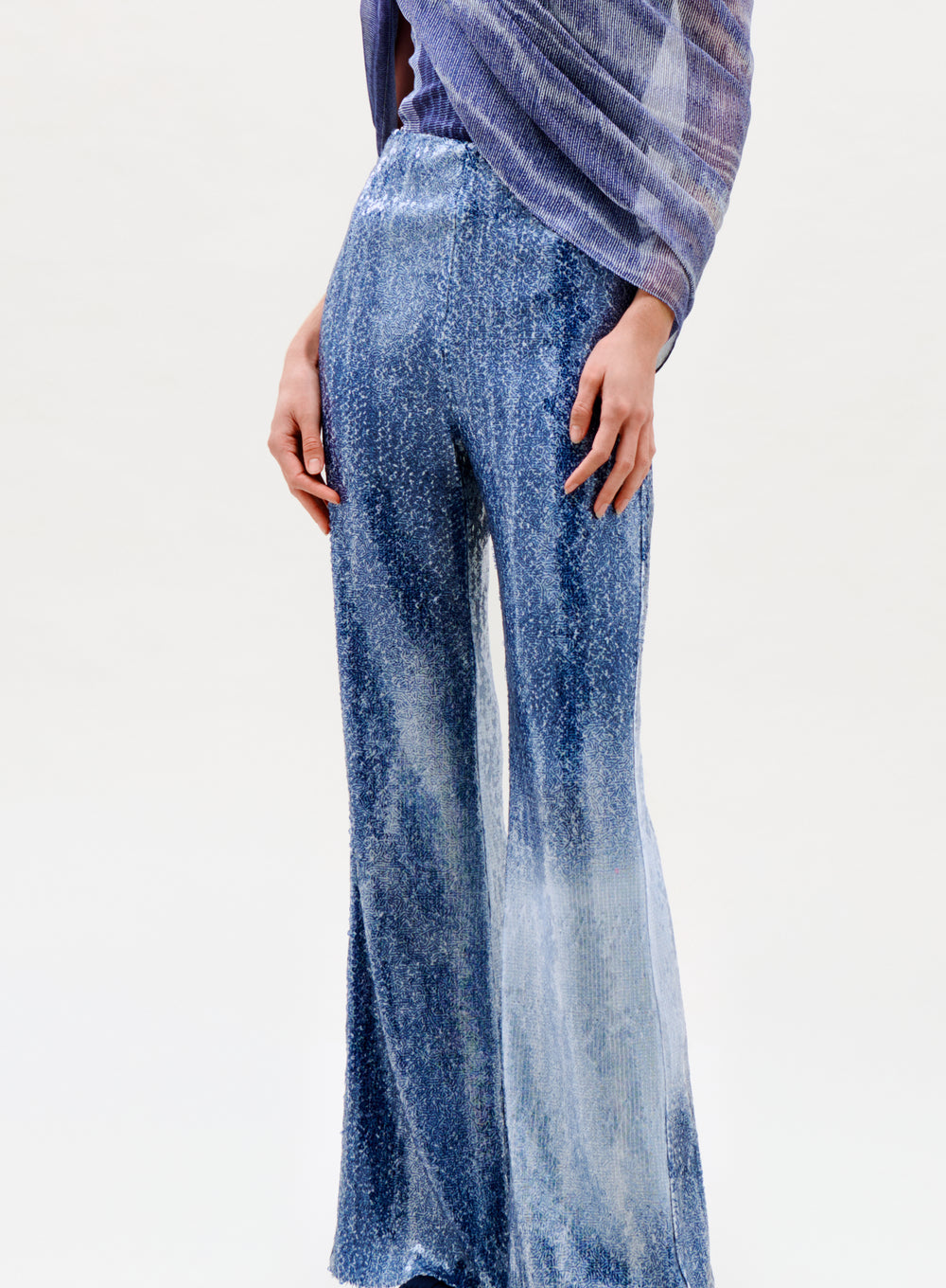 A person wears the Avellino Pant—blue, wide-leg pants crafted from a soft, textured fabric with a subtle sheen—paired with a matching sheer purple shawl. The background is plain and light-colored.