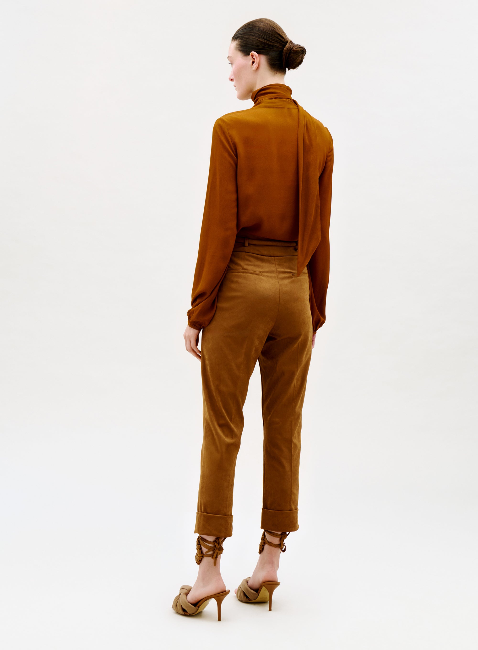 A woman faces away, wearing a long-sleeve brown blouse with a scarf detail, the Beryl Pant with rolled cuffs, and tan heeled mules. Her dark hair is in a neat bun against a plain light background.