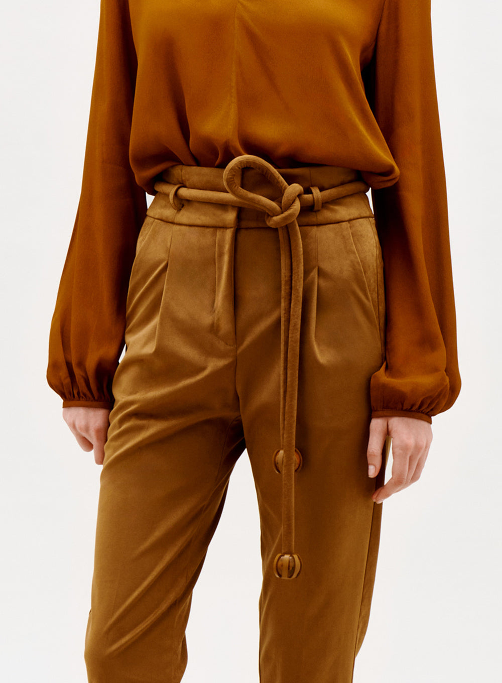 A person wears the Beryl Pant—high-waisted brown trousers with a matching fabric belt—and a long-sleeved rust blouse. The image is cropped above the shoulders and below the knees.