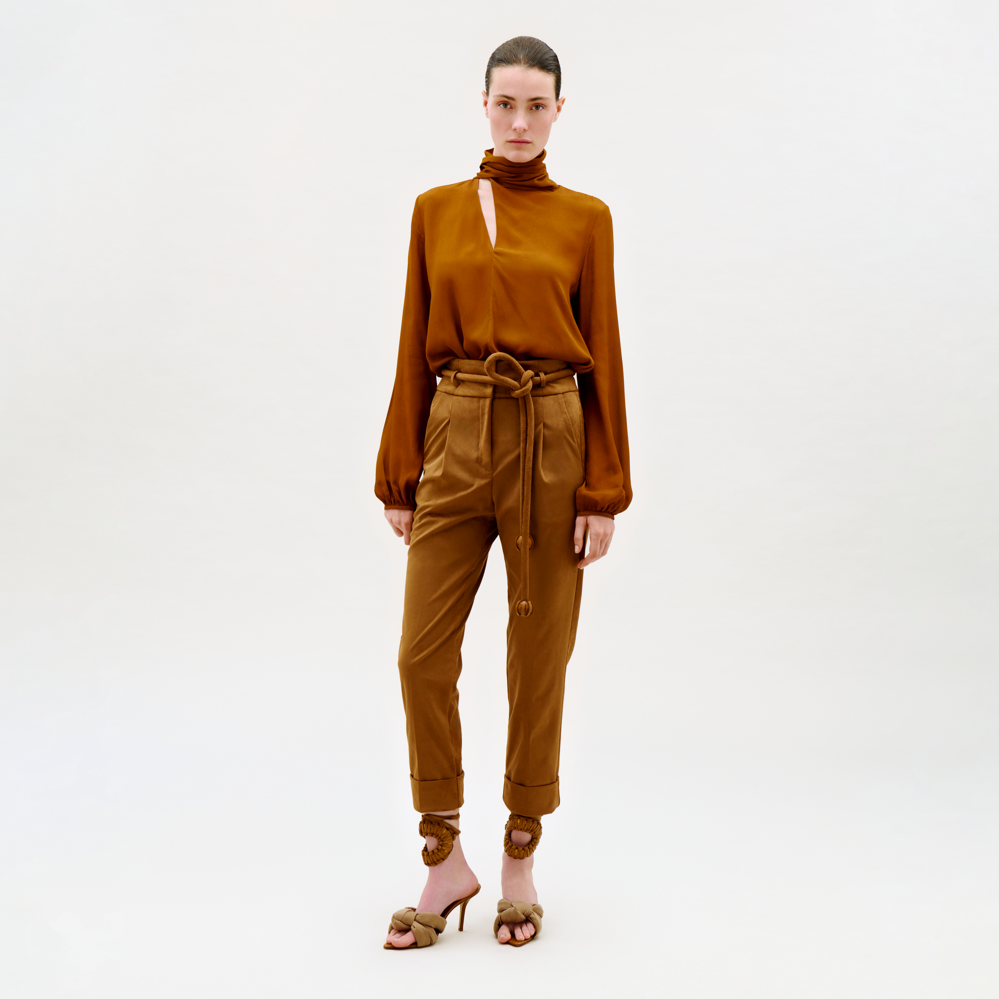A person stands against a plain light background wearing a rust-colored high-neck blouse with a front cutout, the tan Beryl Pant with a belt and high waist, and brown heeled sandals with ankle ties.