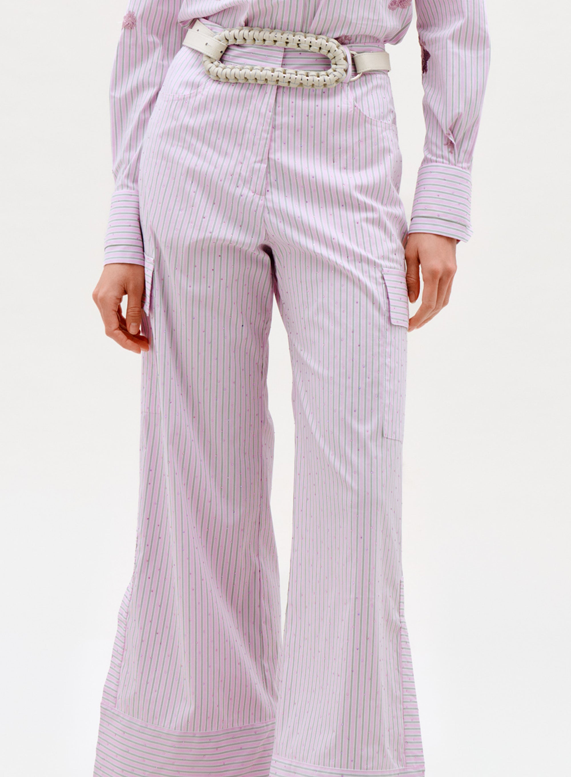 Wearing Falak Pant: high-waisted, wide-leg, light pink pinstriped pants paired with a matching shirt and white braided belt; shown from mid-chest to feet against a plain background.