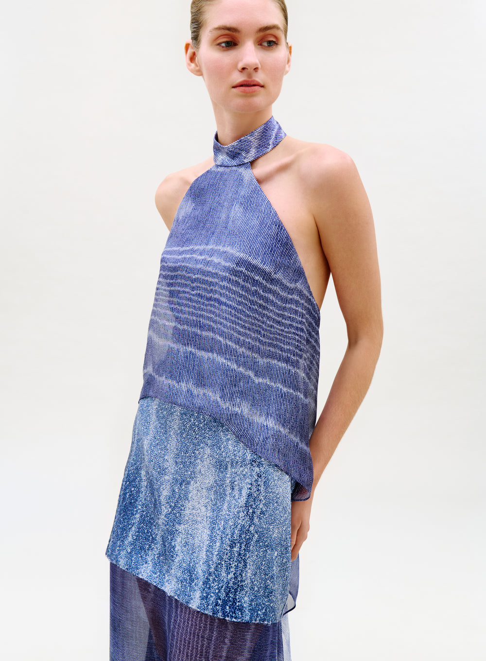 A woman models the Flo Top, a sleeveless high-neck blue top with semi-sheer layered textures and patterns. She stands against a plain light background, looking left with her hand by her side.