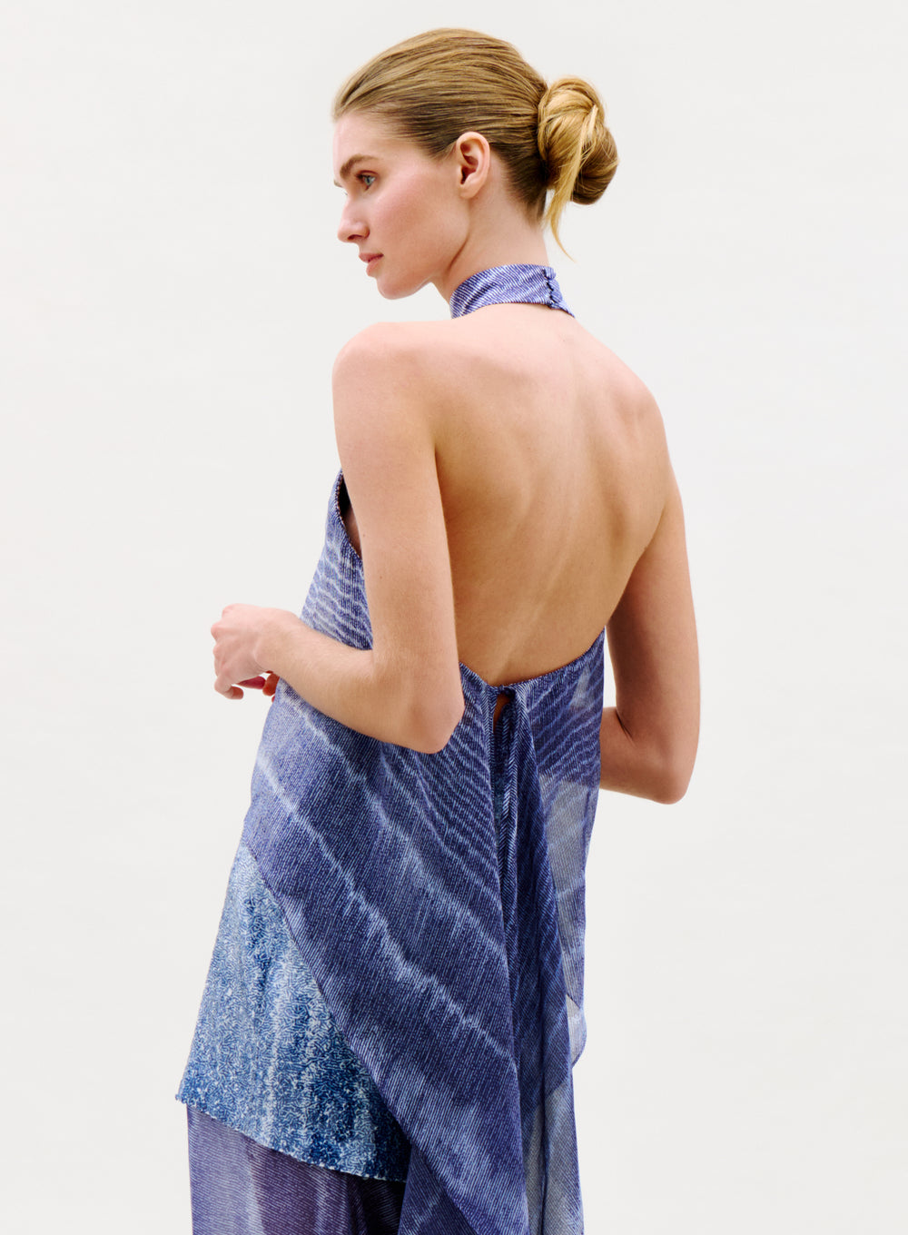A woman with blonde hair in a sleek bun models the sleeveless, backless Flo Top in blue with a textured pattern, standing against a plain light background, seen from the side and slightly from behind.