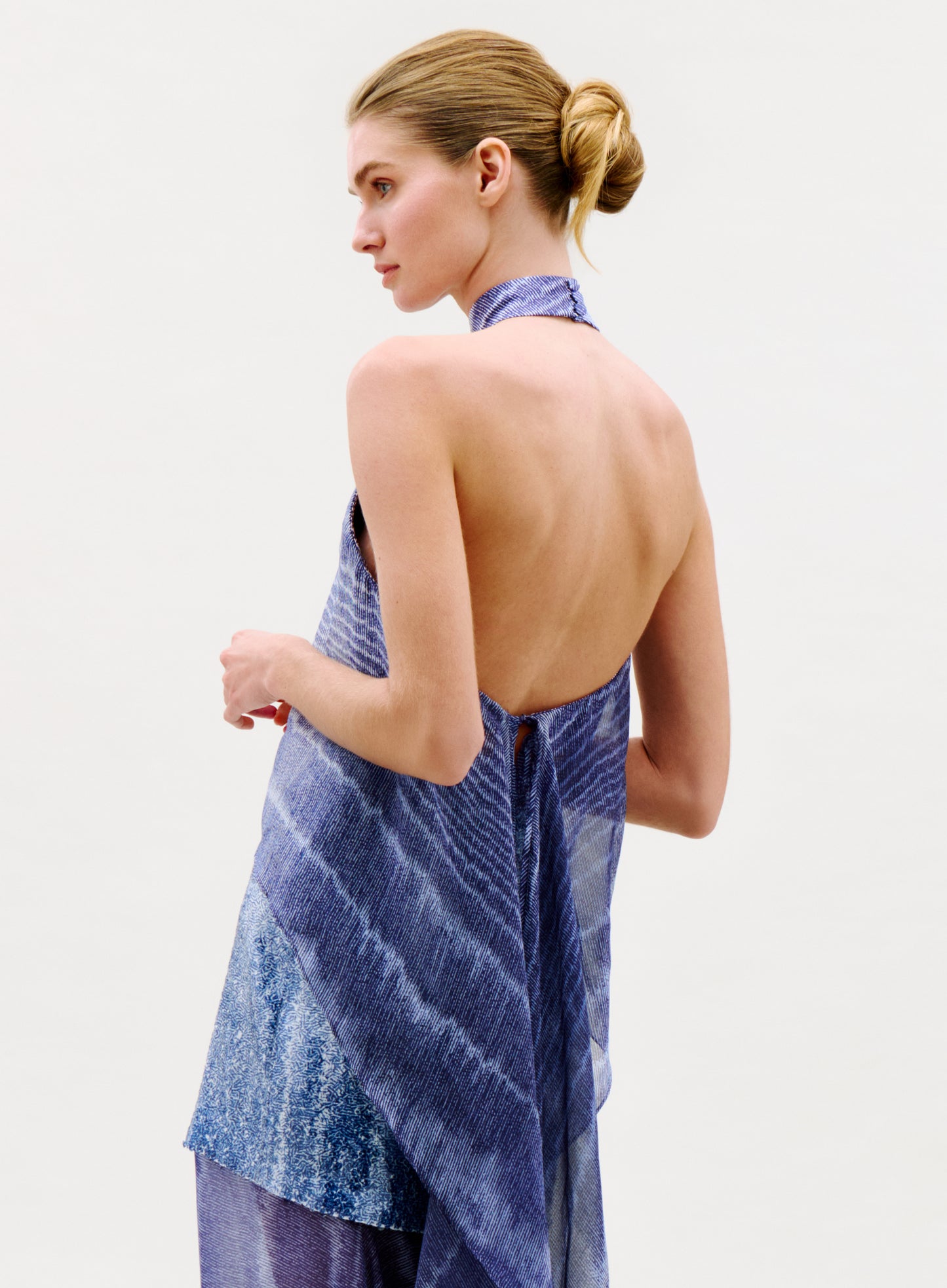 A woman with blonde hair in a sleek bun models the sleeveless, backless Flo Top in blue with a textured pattern, standing against a plain light background, seen from the side and slightly from behind.