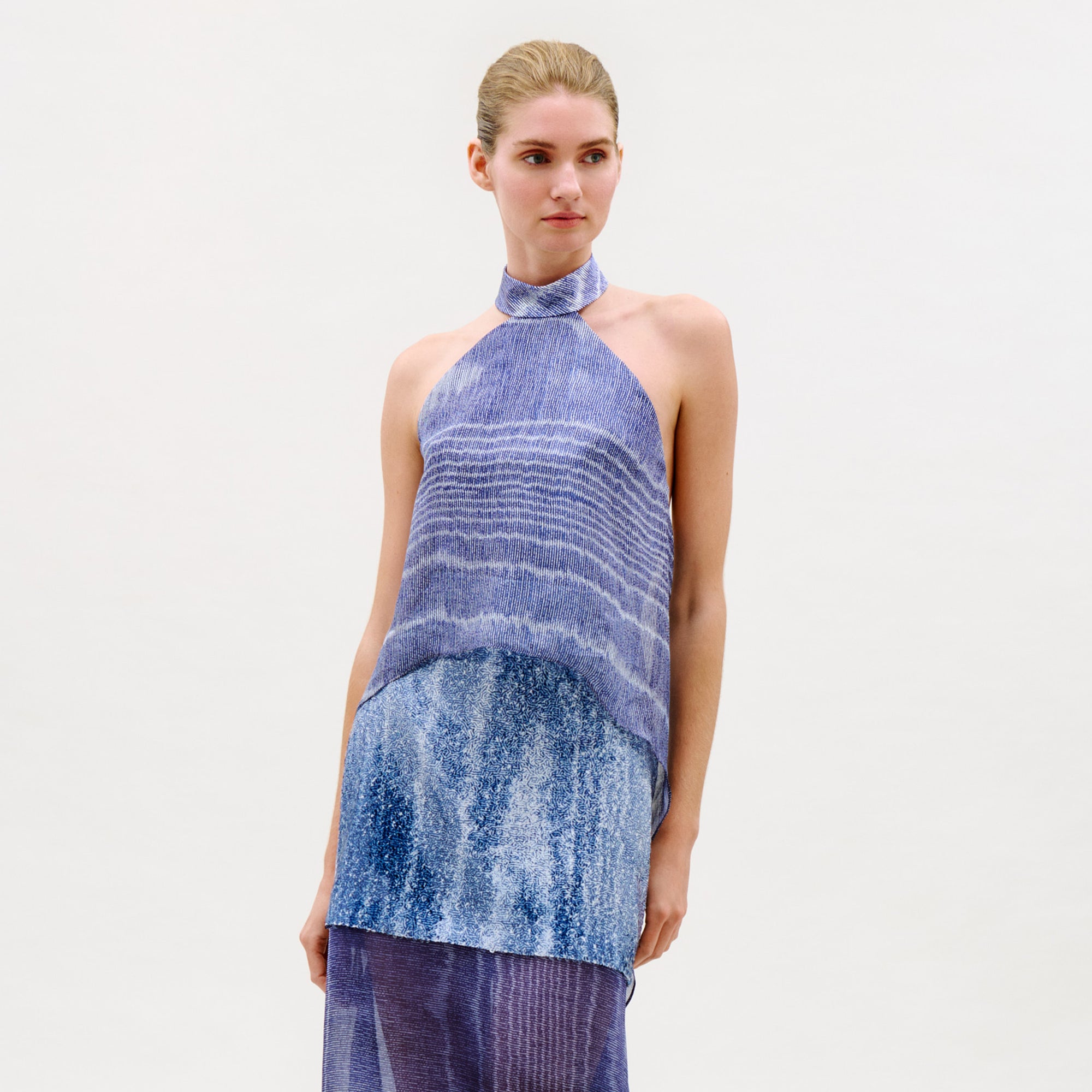 A woman with blonde hair in a low bun wears the sleeveless, halter-neck Flo Top featuring blue and purple gradient horizontal stripes, paired with a matching skirt against a plain light background.