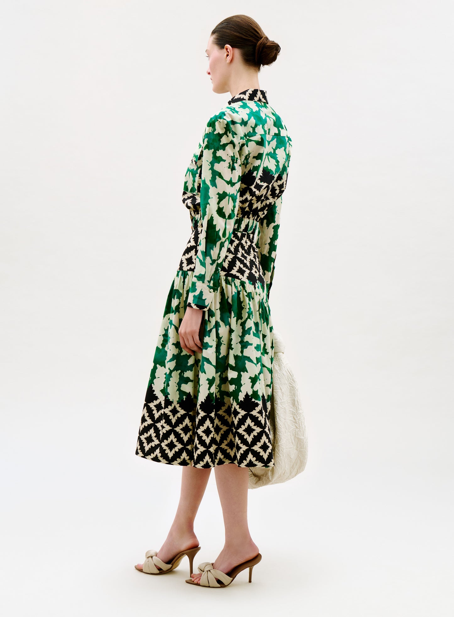 A woman stands in profile against a white background, wearing the Libby Skirt with a long-sleeved, green, black, and white patterned top, beige heeled sandals, and holding a textured white bag.